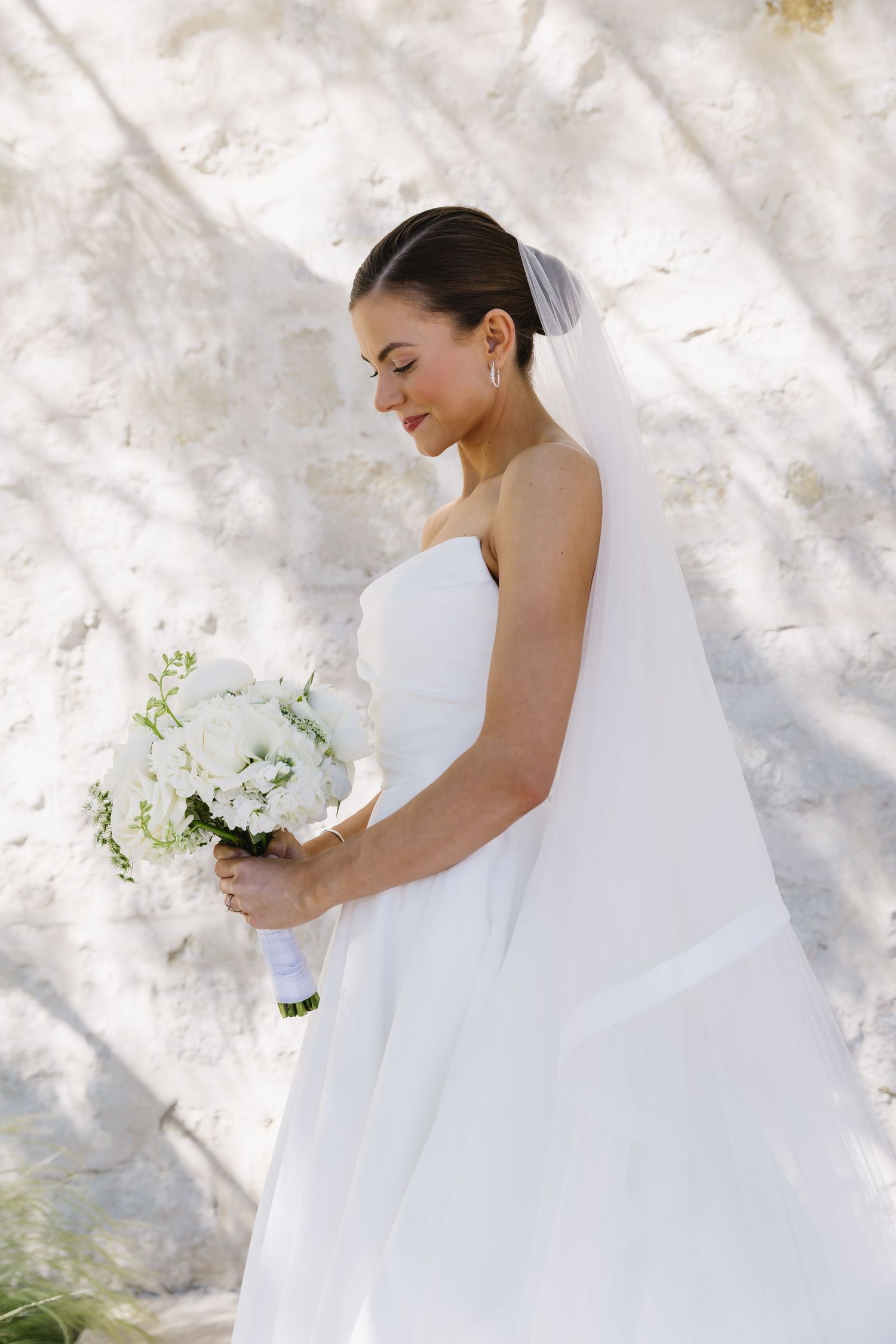 A bride in a flowing white wedding dress holds a white bouquet against a light colored textured wall background.