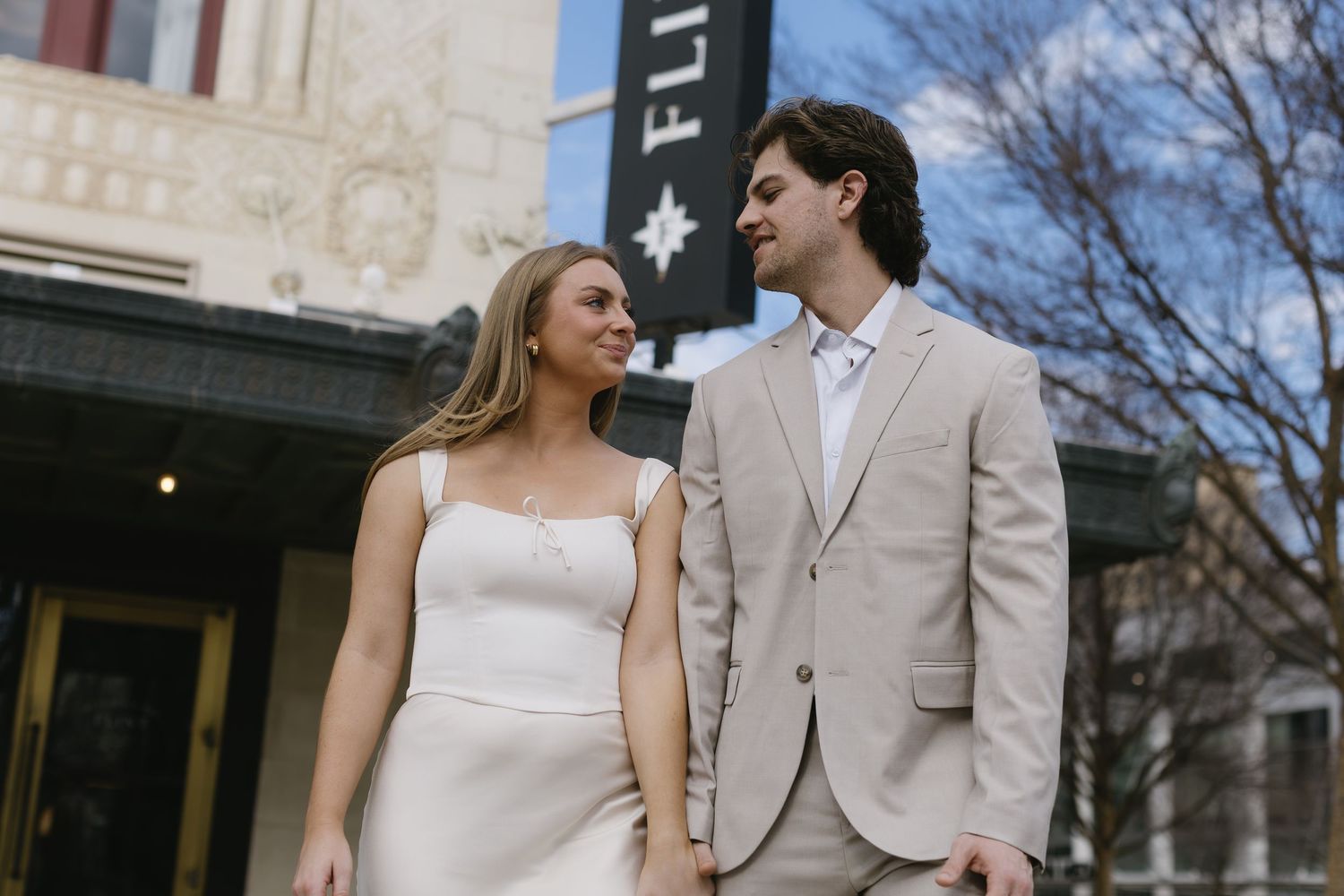 Couple in formal wear walking together outside a city building on a sunny day.