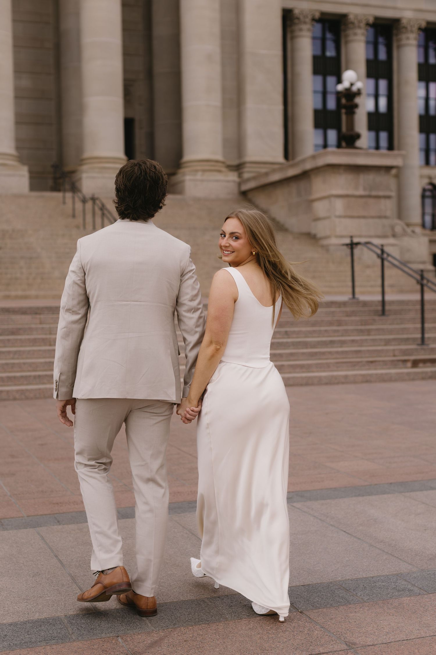 A couple in wedding attire walking up grand stone steps, with the person in a light suit leading the way.
