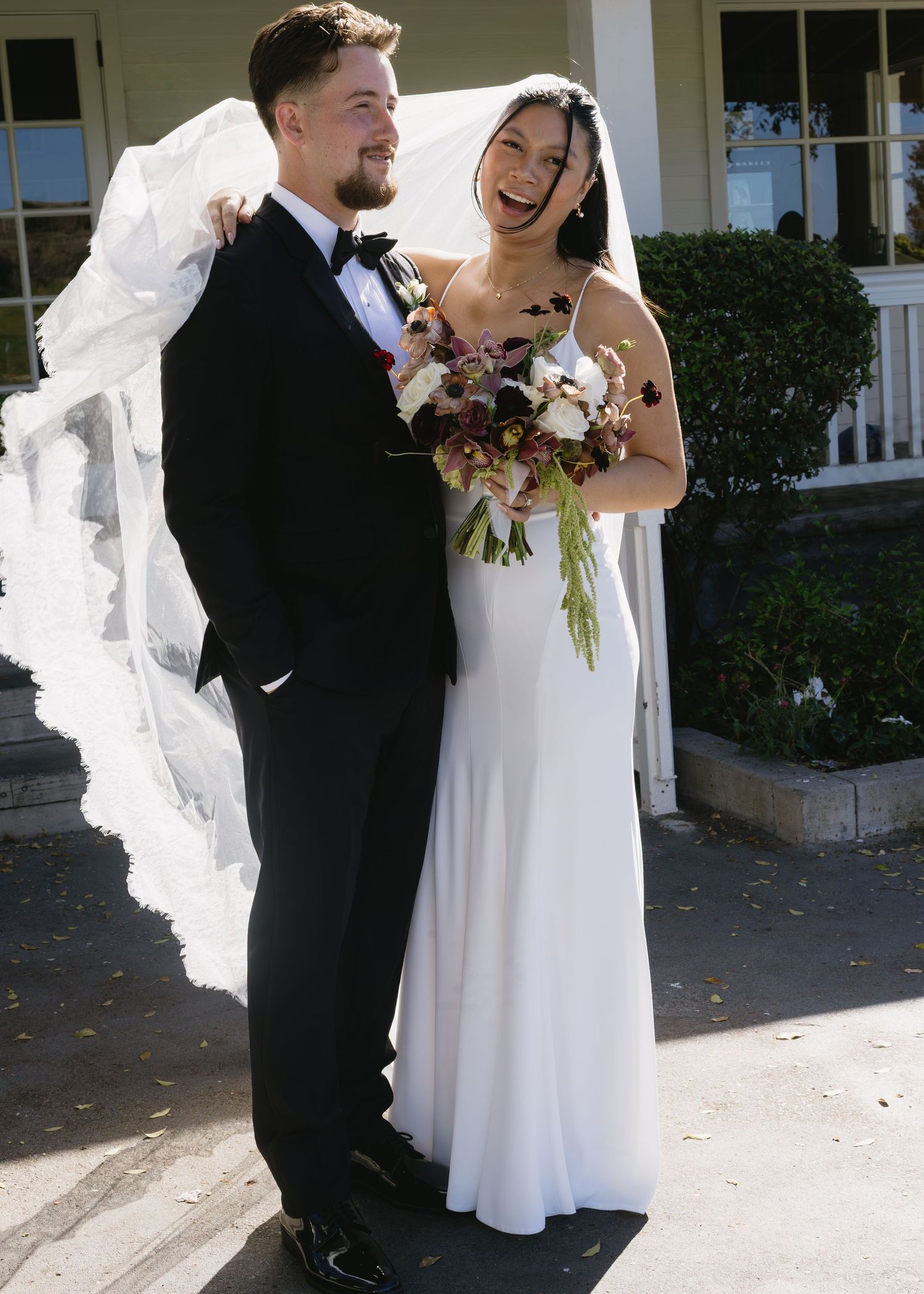 Happy newlyweds share a laugh while holding burgundy and white floral bouquet.