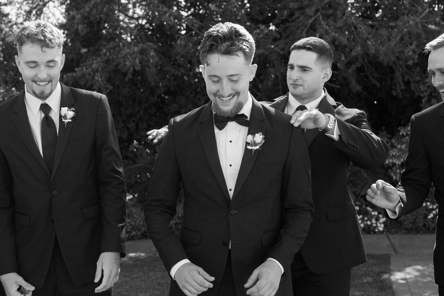 Groomsmen in black tuxedos share candid moments during outdoor wedding photography.