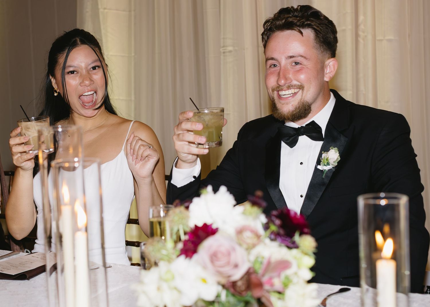 A couple shares a joyful moment at their wedding reception dinner table with champagne glasses.