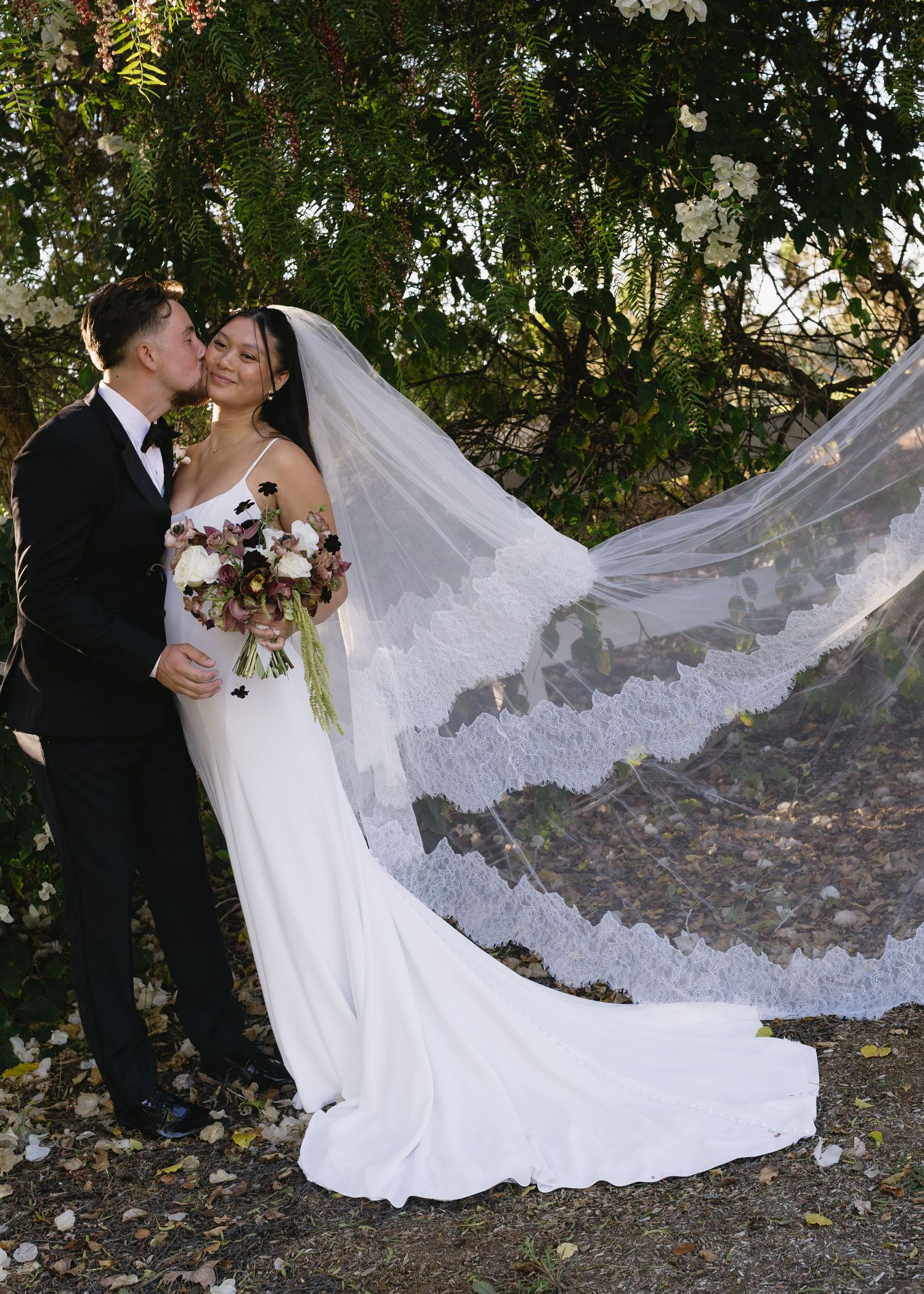 Wedding couple poses for romantic photos with dramatic flowing veil on garden path.