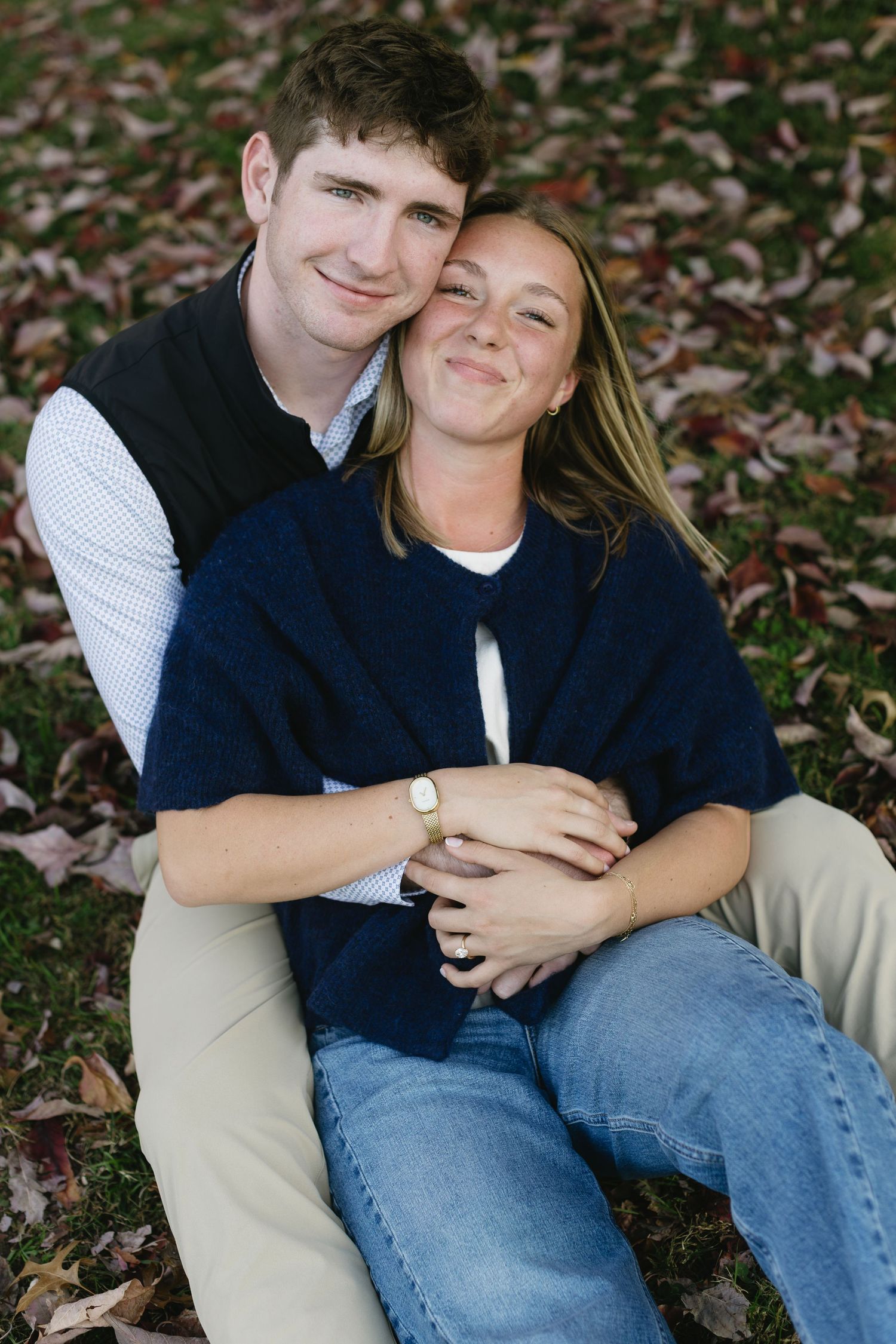 A couple sits together on fallen autumn leaves sharing an intimate moment in casual attire.