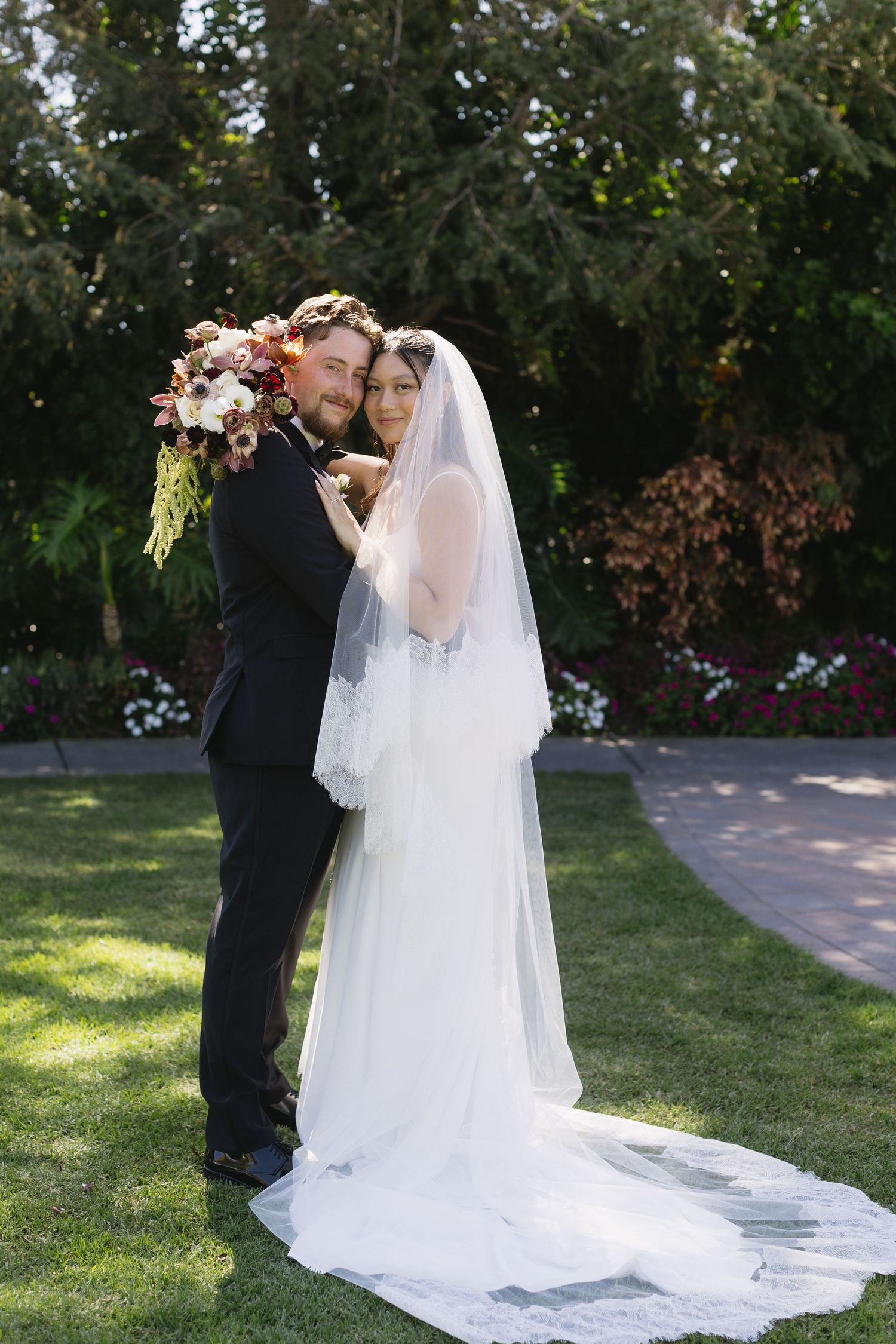 A romantic wedding couple shares a tender kiss in a sunlit garden setting with a flowing white veil trailing behind them.
