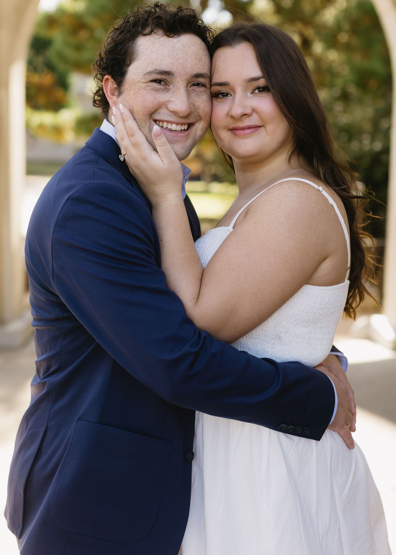 A couple in wedding attire embraces and smiles during an outdoor photo session under a covered walkway.