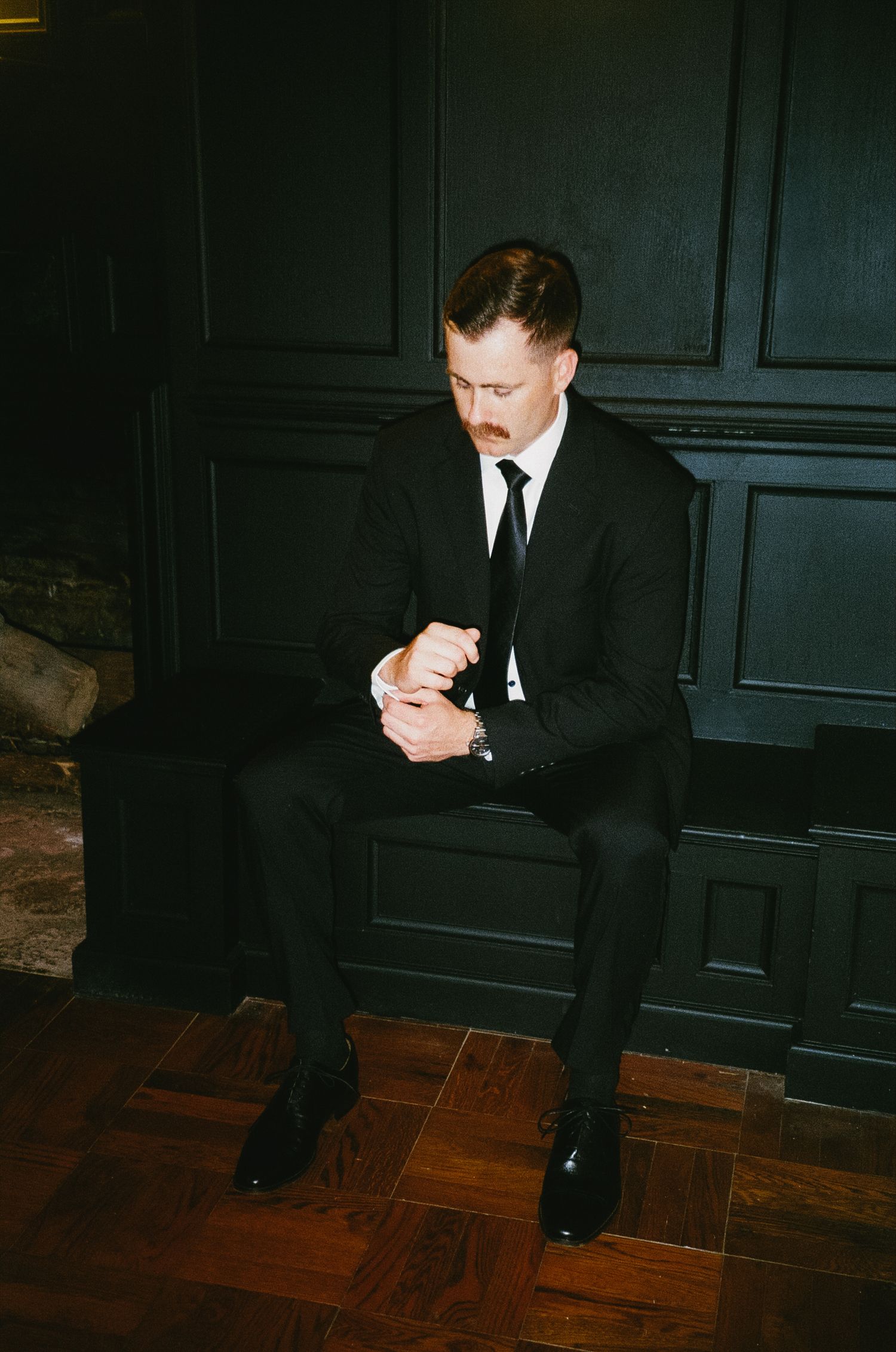 A series of moody shots showing someone in a black suit seated on a hardwood floor against a dark wall.