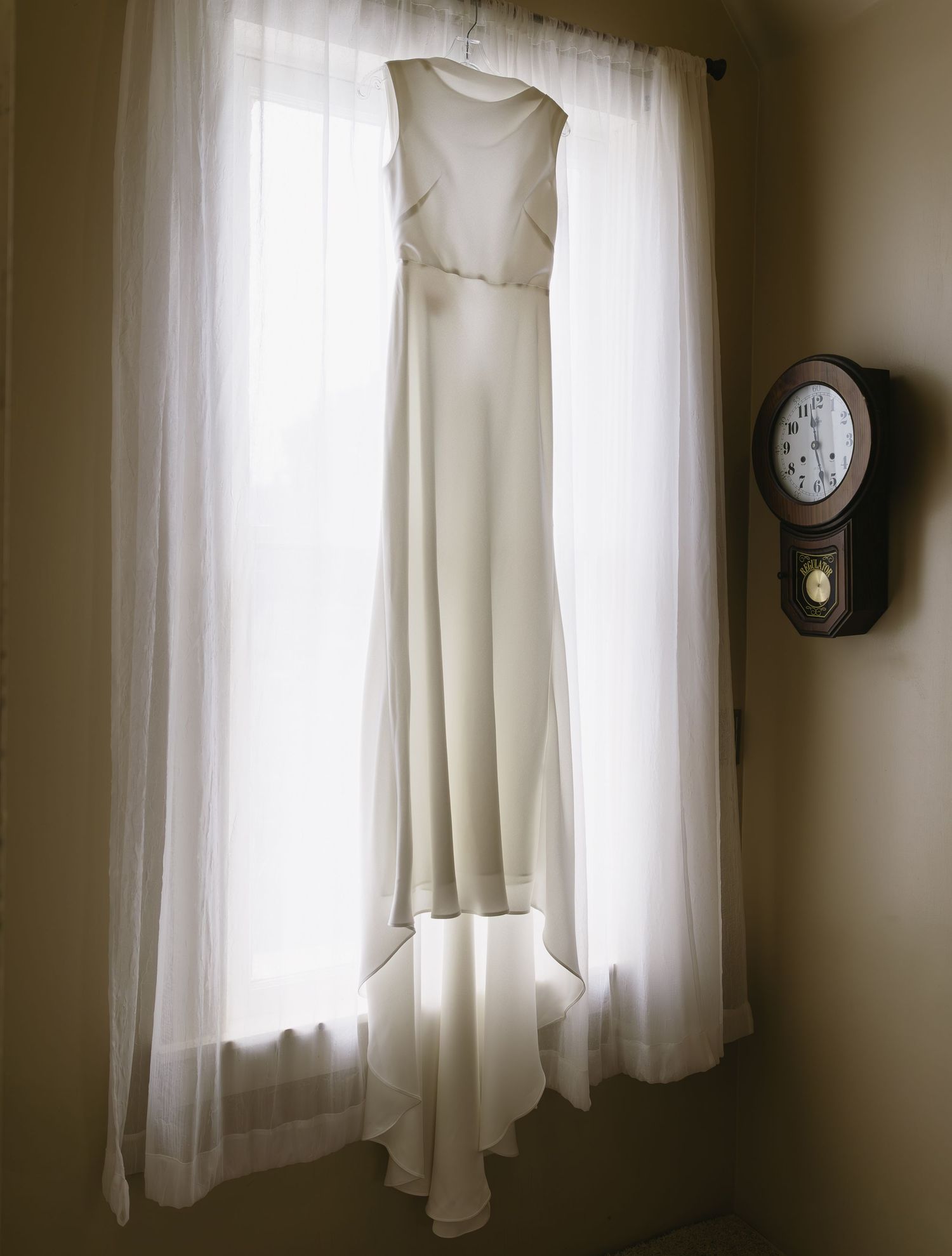 White wedding dress hangs near windows with sheer curtains in natural light creating an ethereal atmosphere.