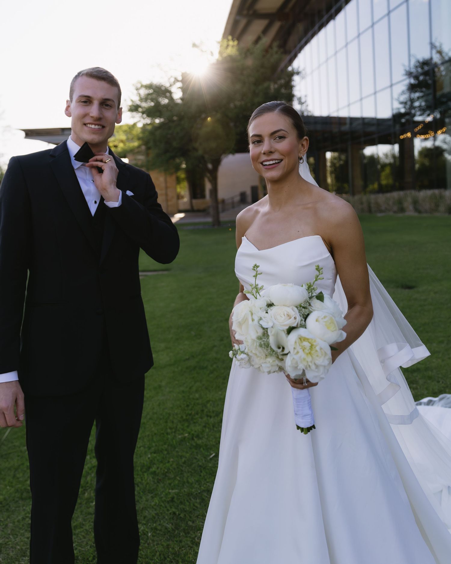 Bride in an elegant white wedding gown holds a white floral bouquet against a scenic outdoor backdrop.
