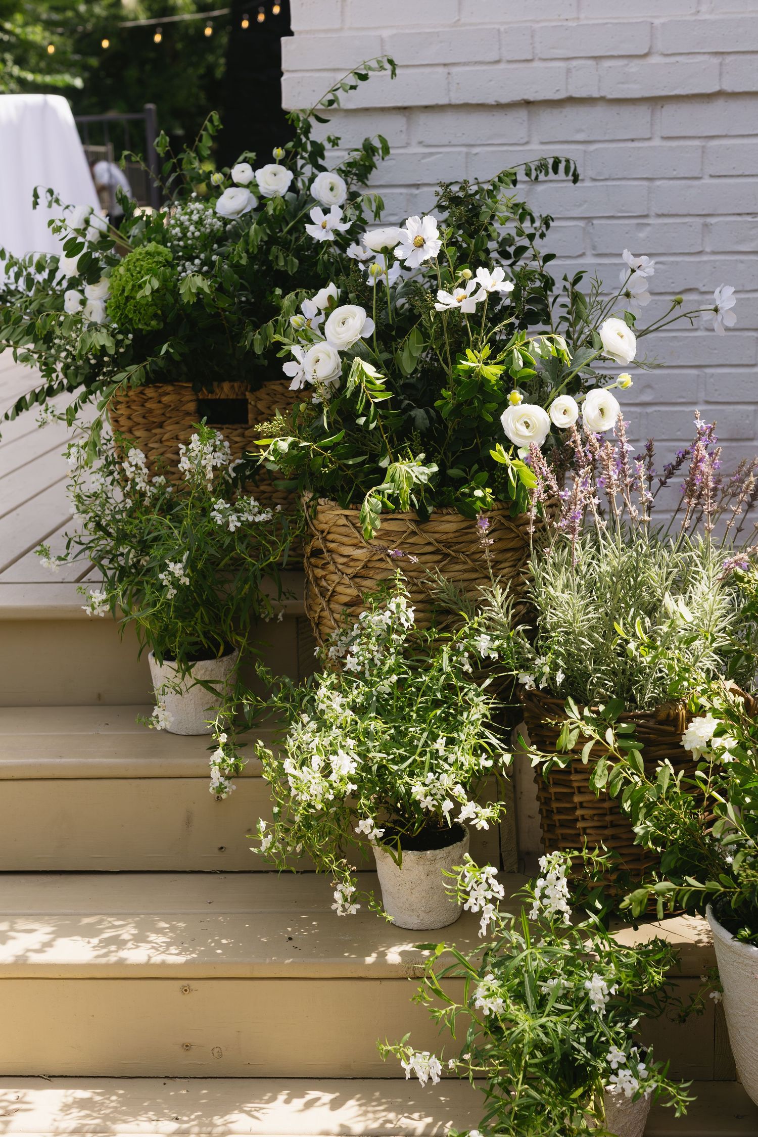 White flowers and greenery decorate an outdoor garden wall with hanging string lights.