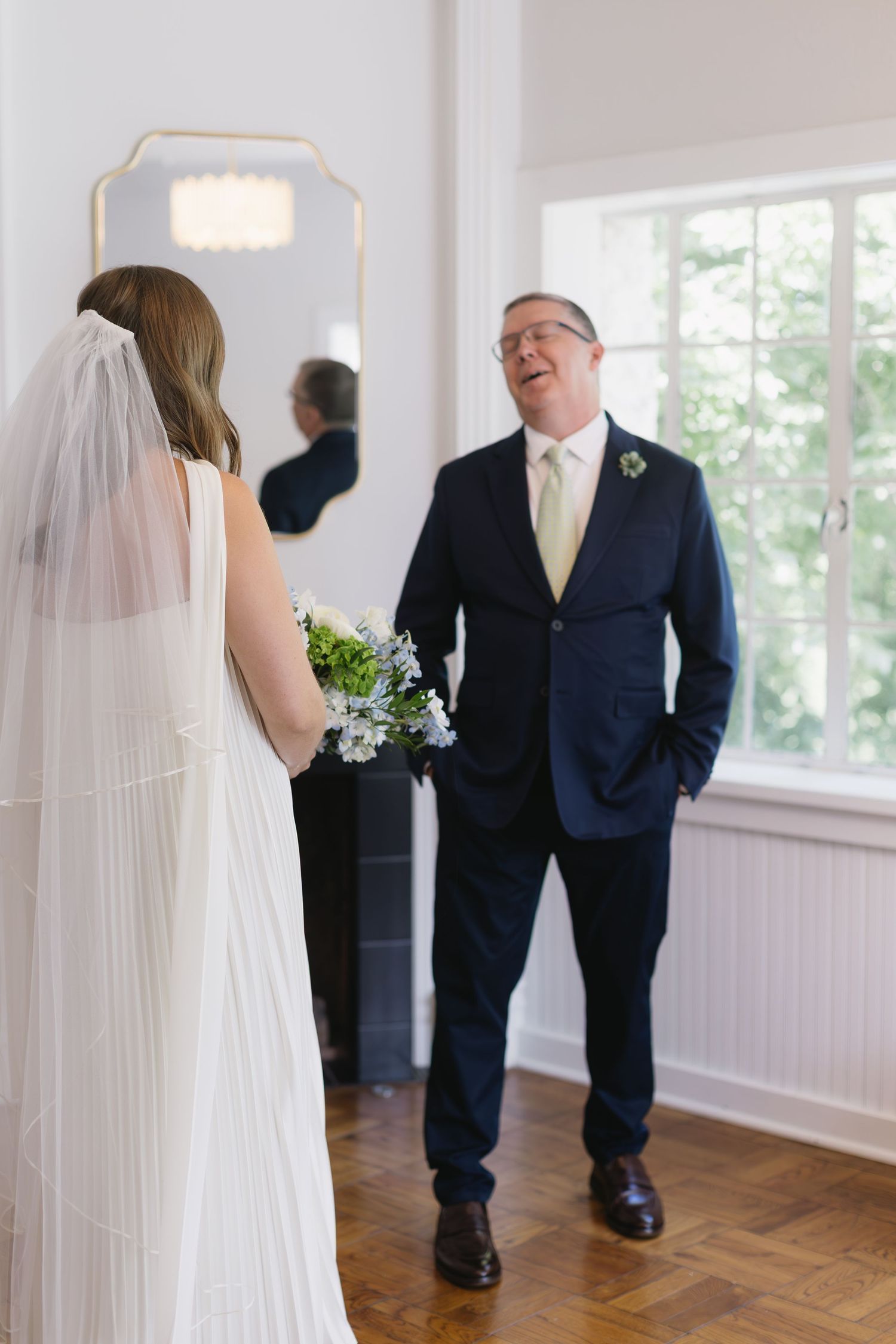 Emotional first look between bride and father in sunlit room with hardwood floors and white walls.