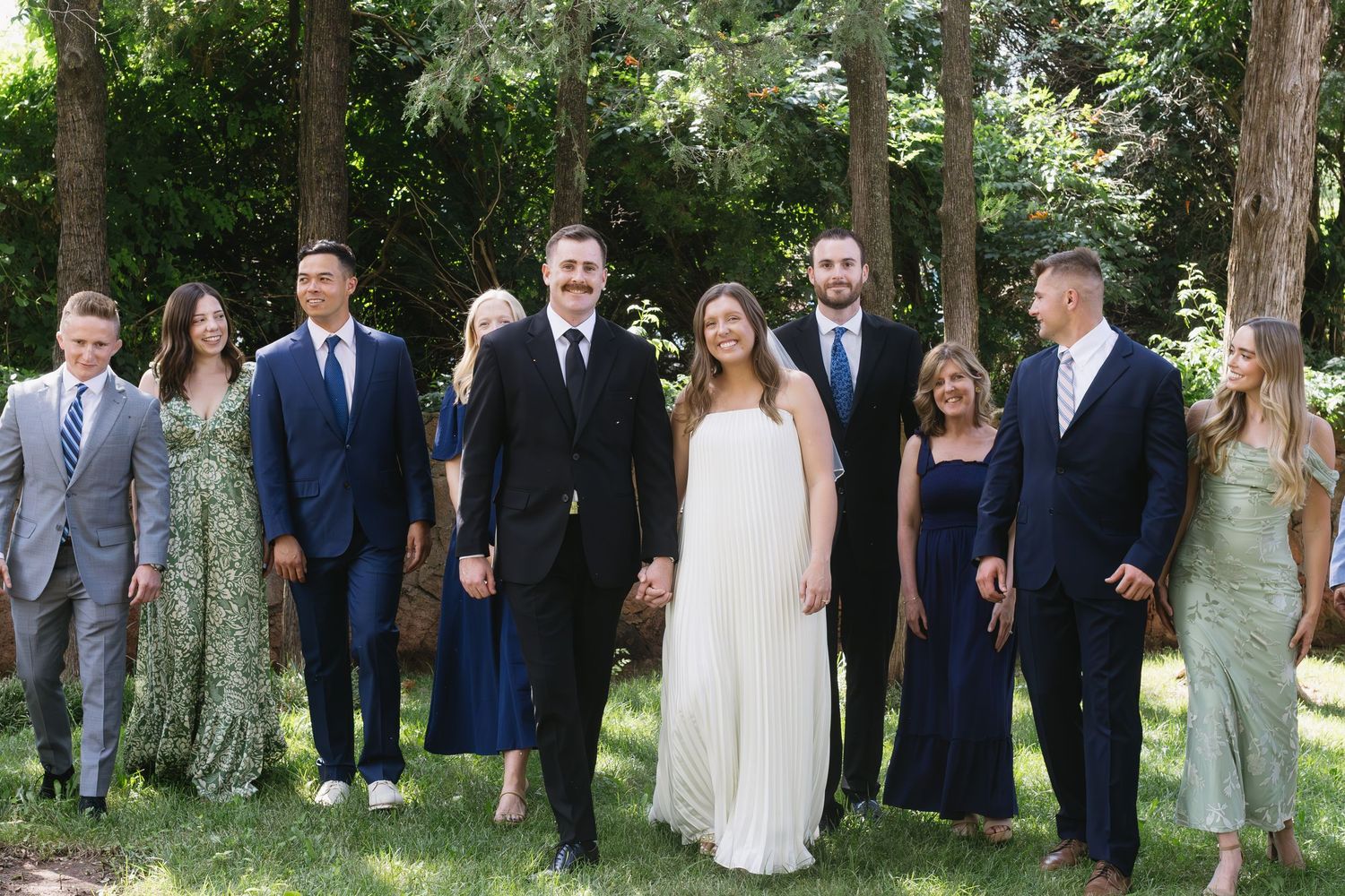 Wedding party posed outdoors among trees wearing coordinated formal attire.