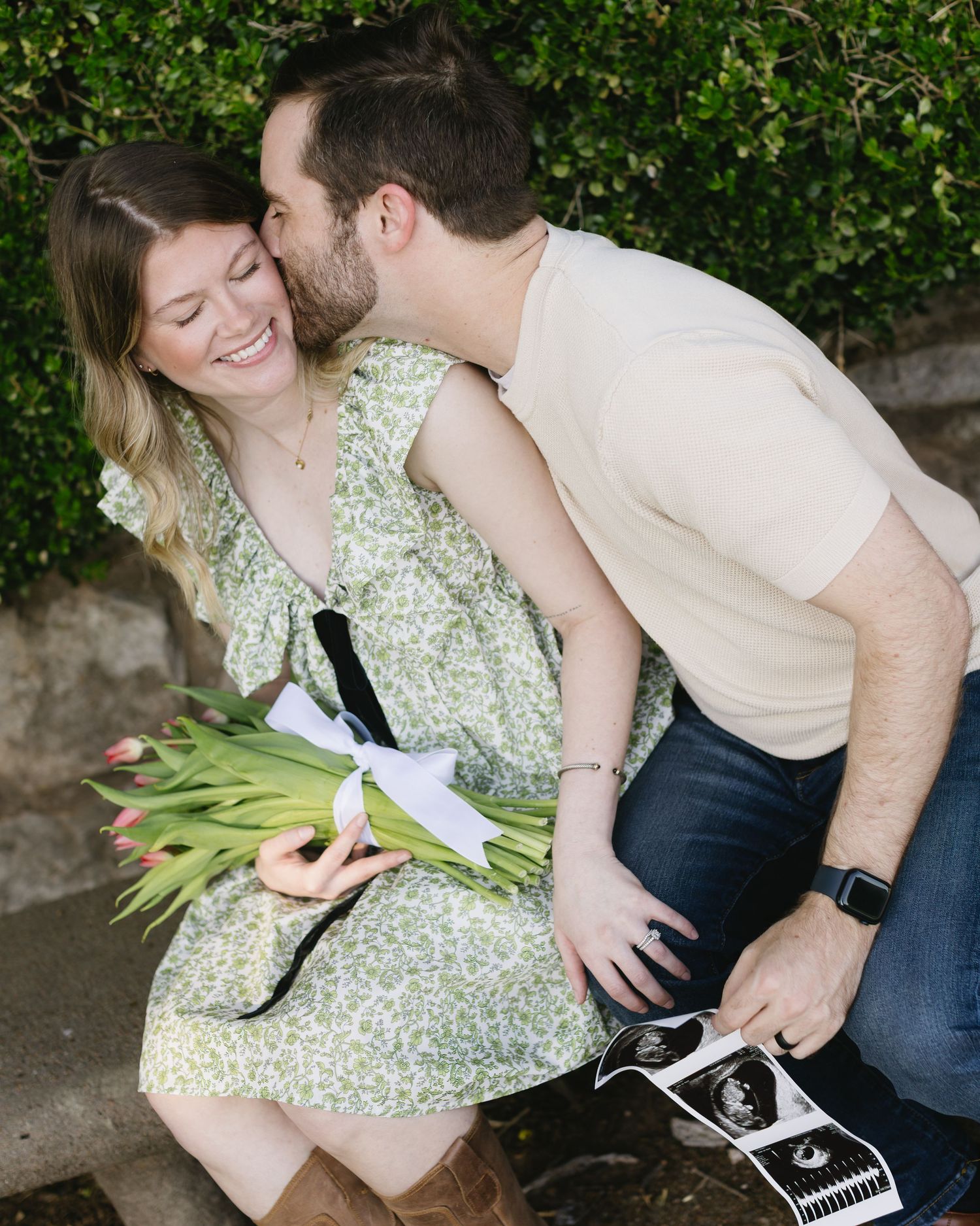 A couple shares an intimate moment while sitting together outdoors on a sunny day near green foliage.