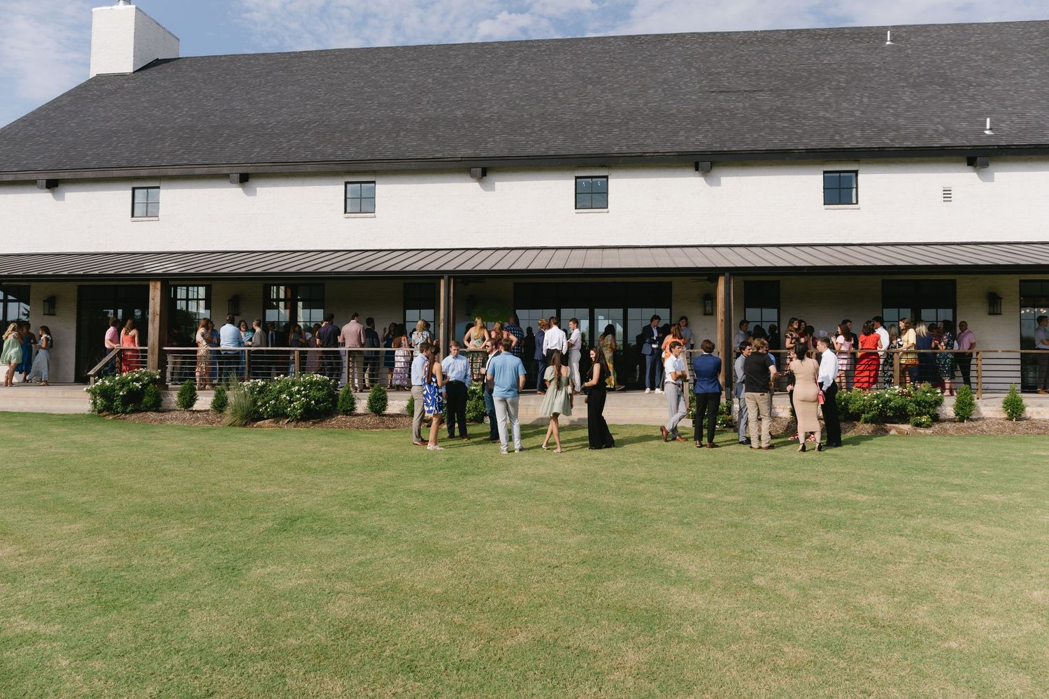 Panoramic view of white barn venue with guests gathered outside on sunny day.