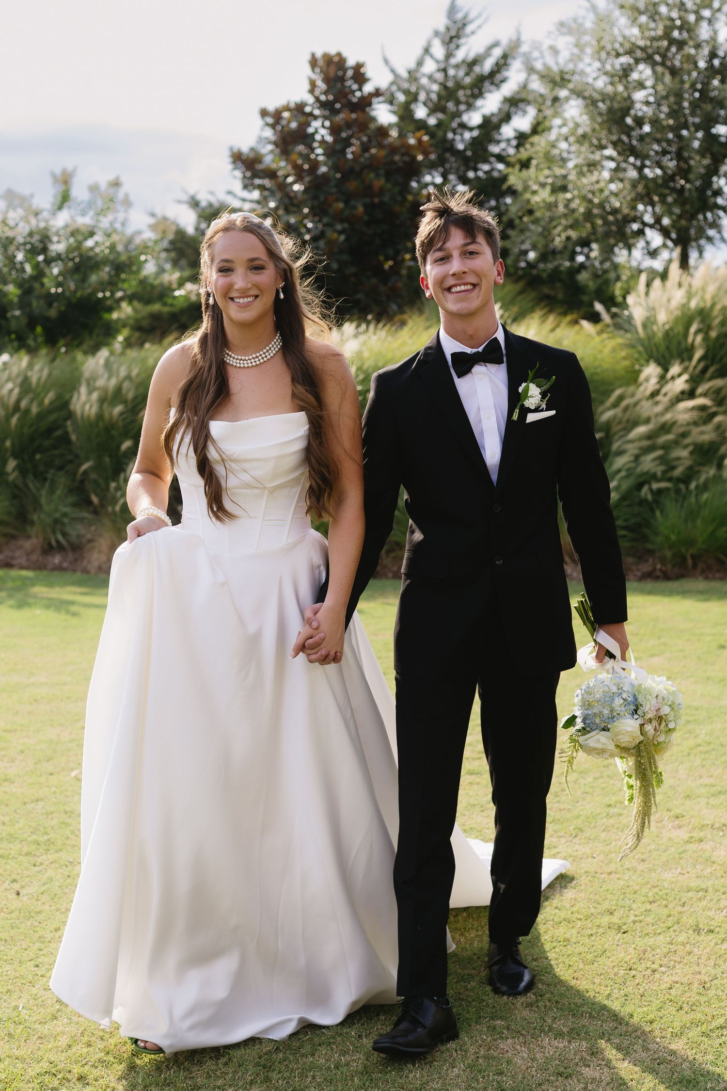 Wedding couple walking together on grassy field in traditional white dress and black tuxedo attire.