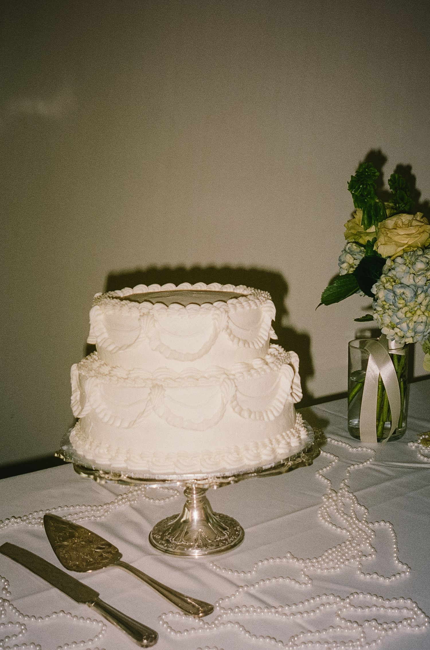 Elegant white wedding cake on silver stand with pearl decorations and floral accents.