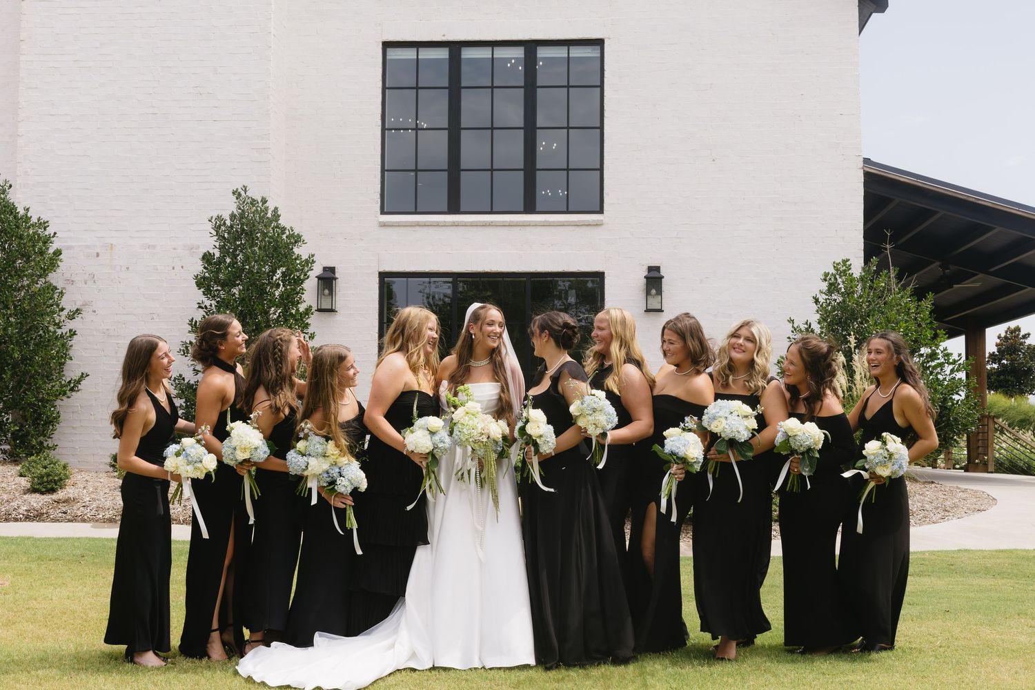 Wedding party in black dresses poses with bride outdoors at modern venue.