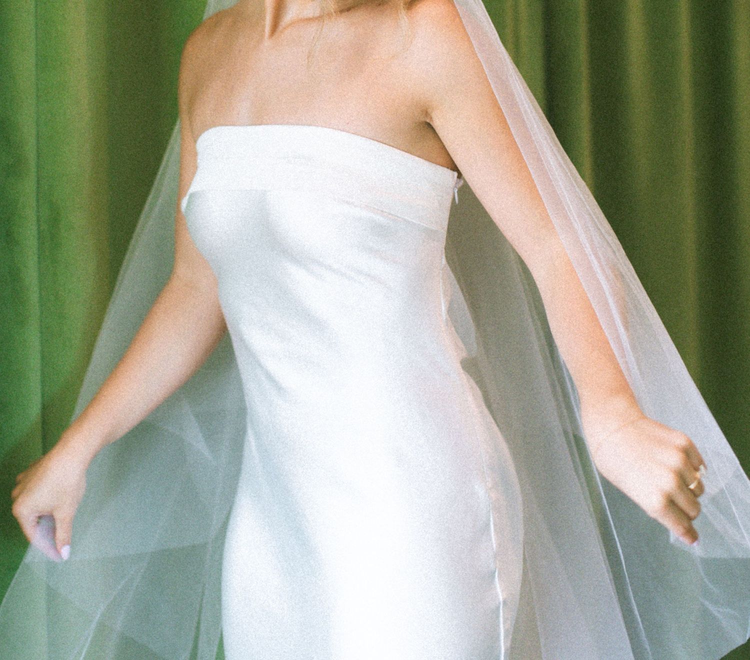 An elegant white wedding dress with flowing veil against a green curtained backdrop.