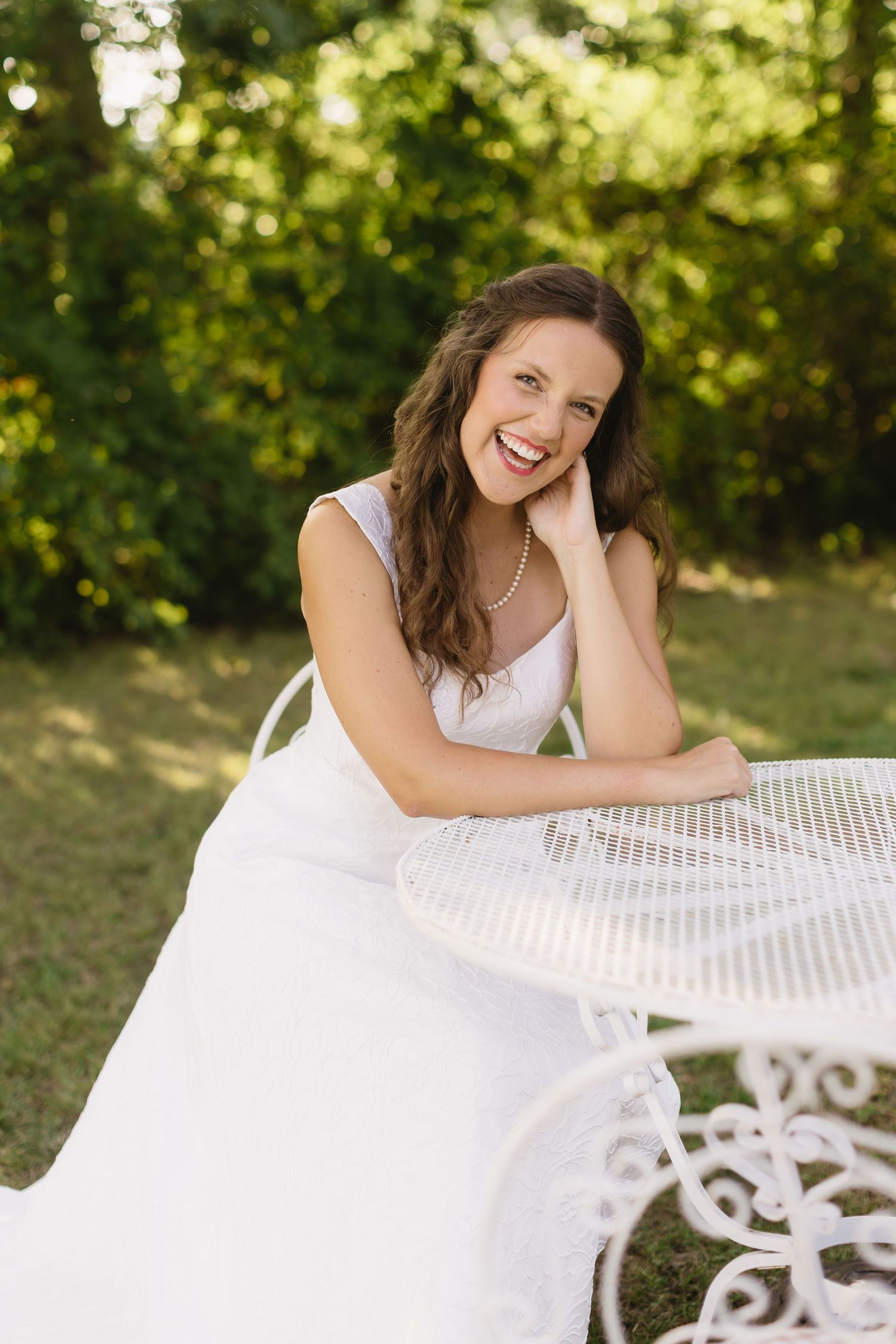 A joyful bride in a white dress sits at an ornate garden table on a sunny day.