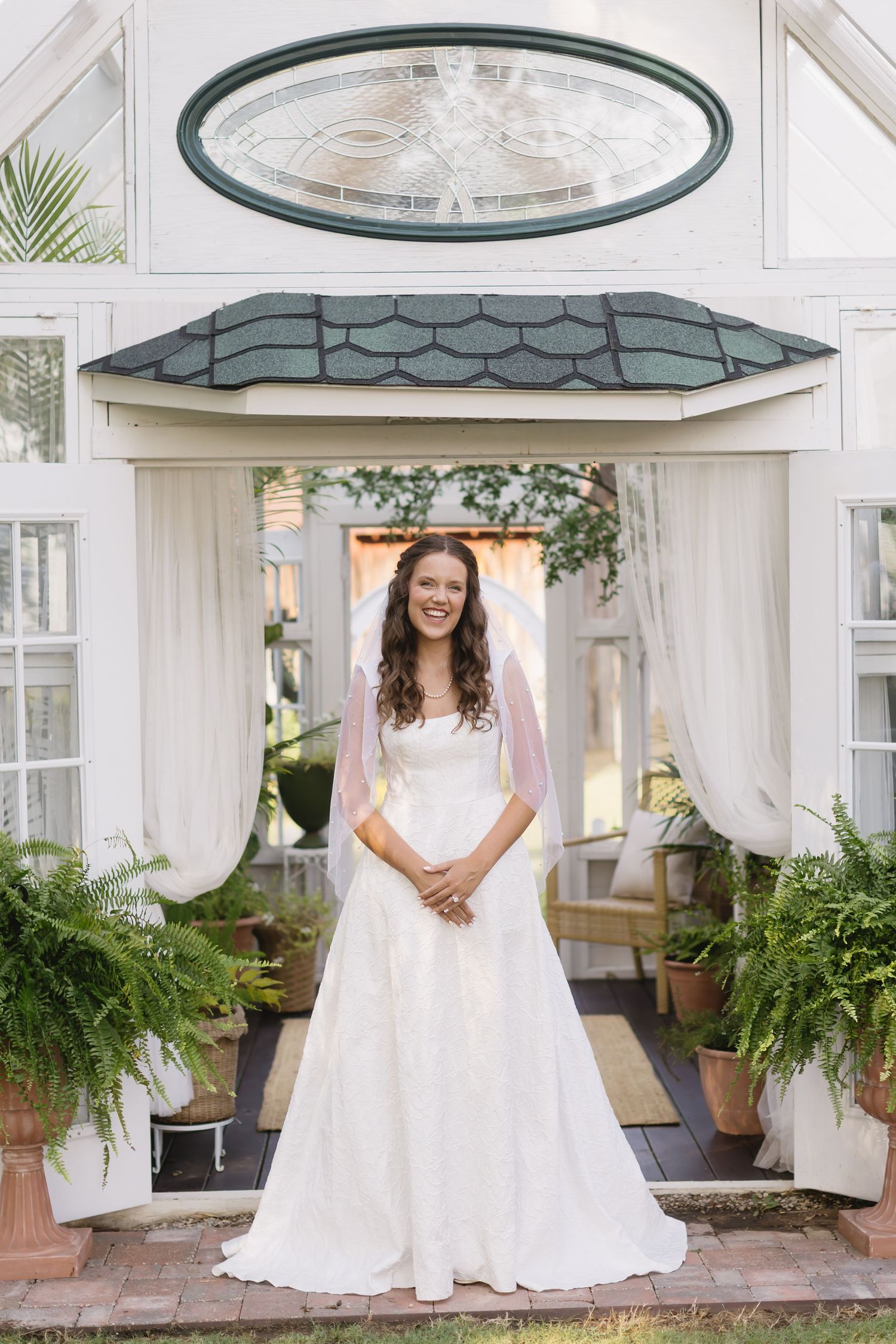 A bride in white wedding dress stands beneath an elegant gazebo with glass ceiling and hanging plants.
