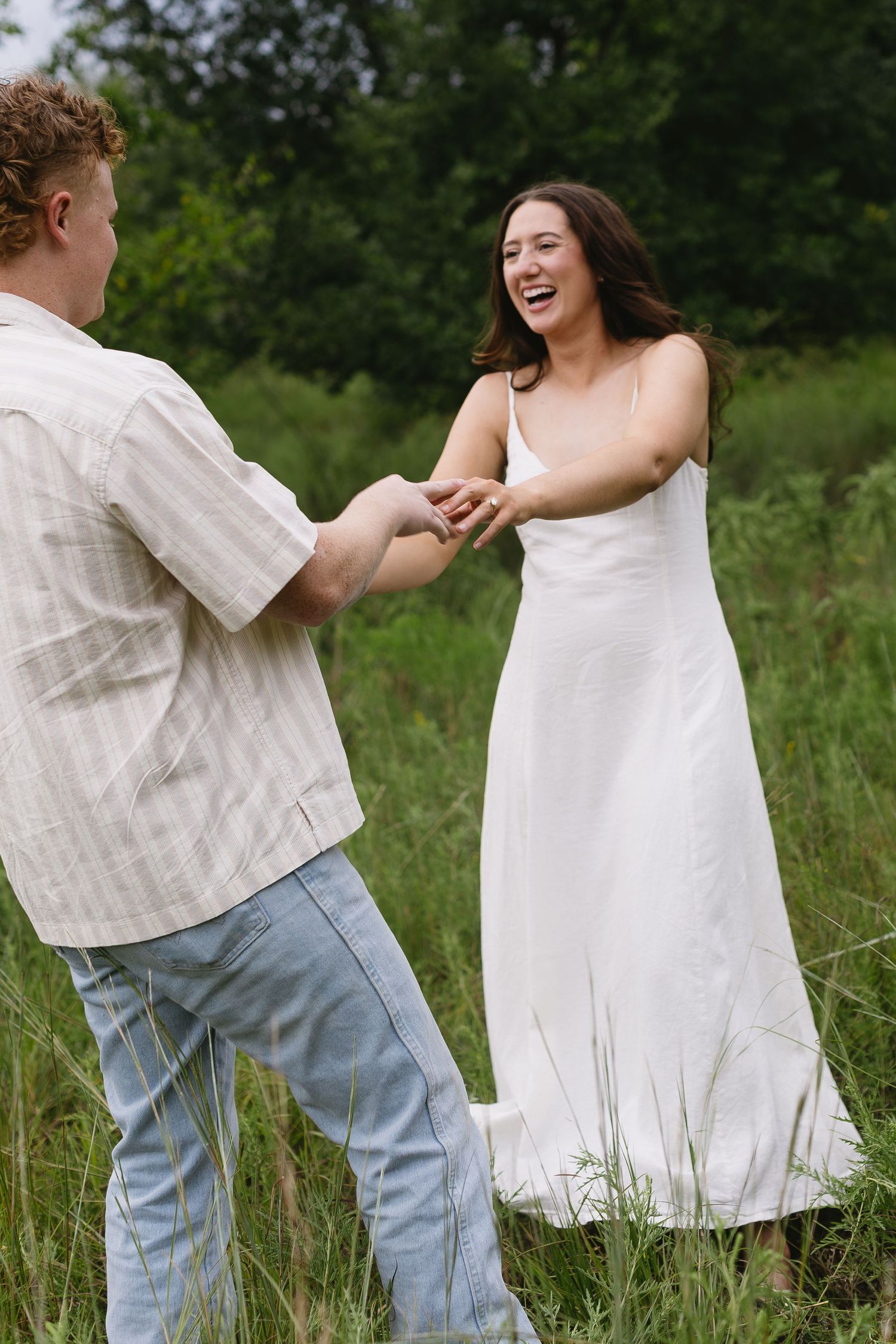 A couple dances playfully in a grassy field, their white and casual outfits flowing with movement.