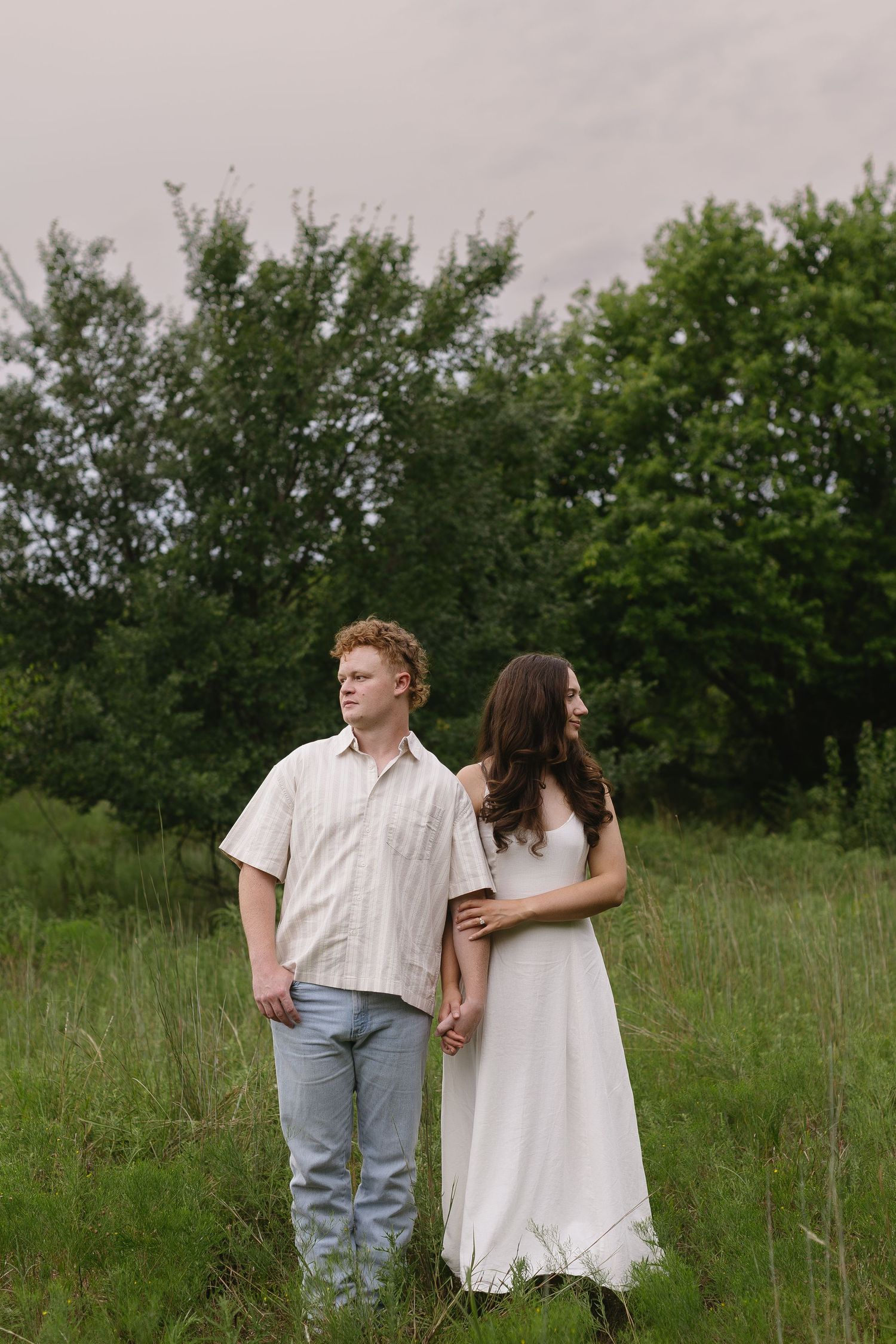 Couple laughing and holding hands while walking through a grassy field in casual summer outfits.