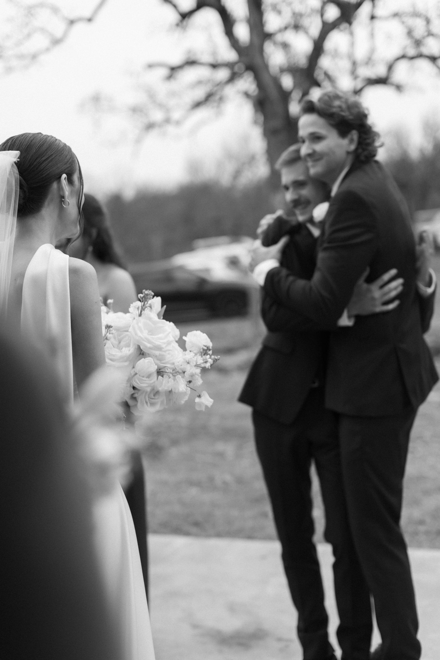Black and white wedding photo showing bride with bouquet watching emotional groomsmen embrace outdoors.