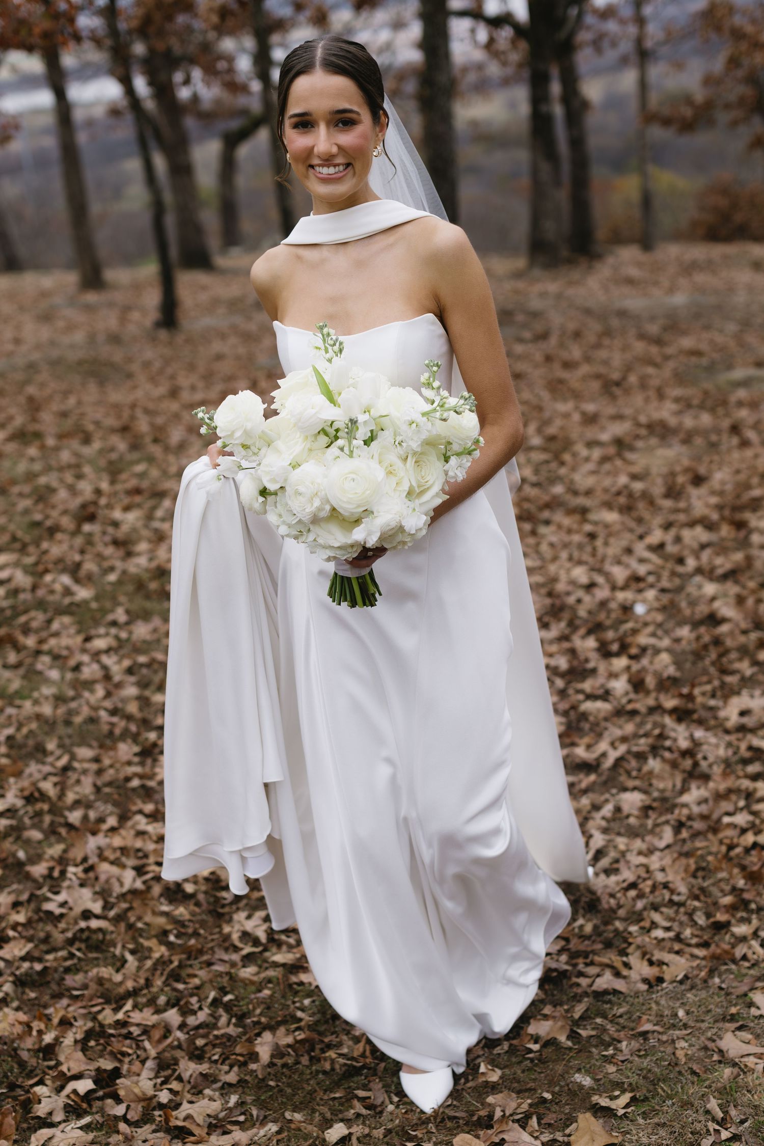 Bride in white strapless gown holding luxurious white bouquet among fall foliage.