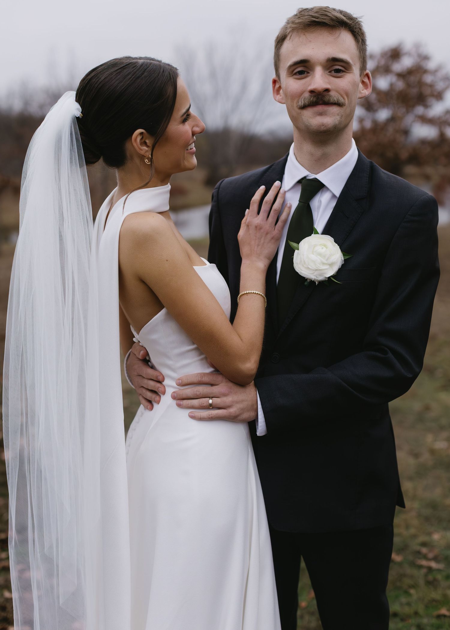 Wedding couple shares tender moment in formal attire against rustic outdoor backdrop.