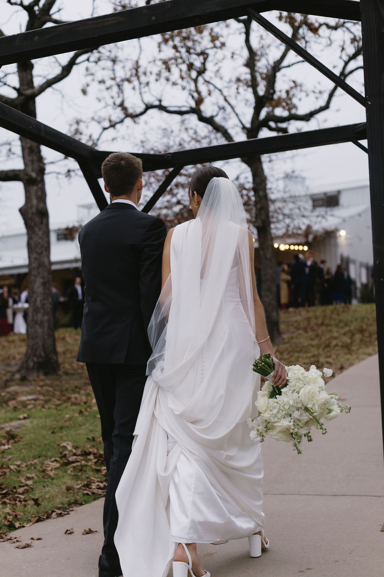 A bride and groom walk along a tree-lined path with her white dress flowing behind them.