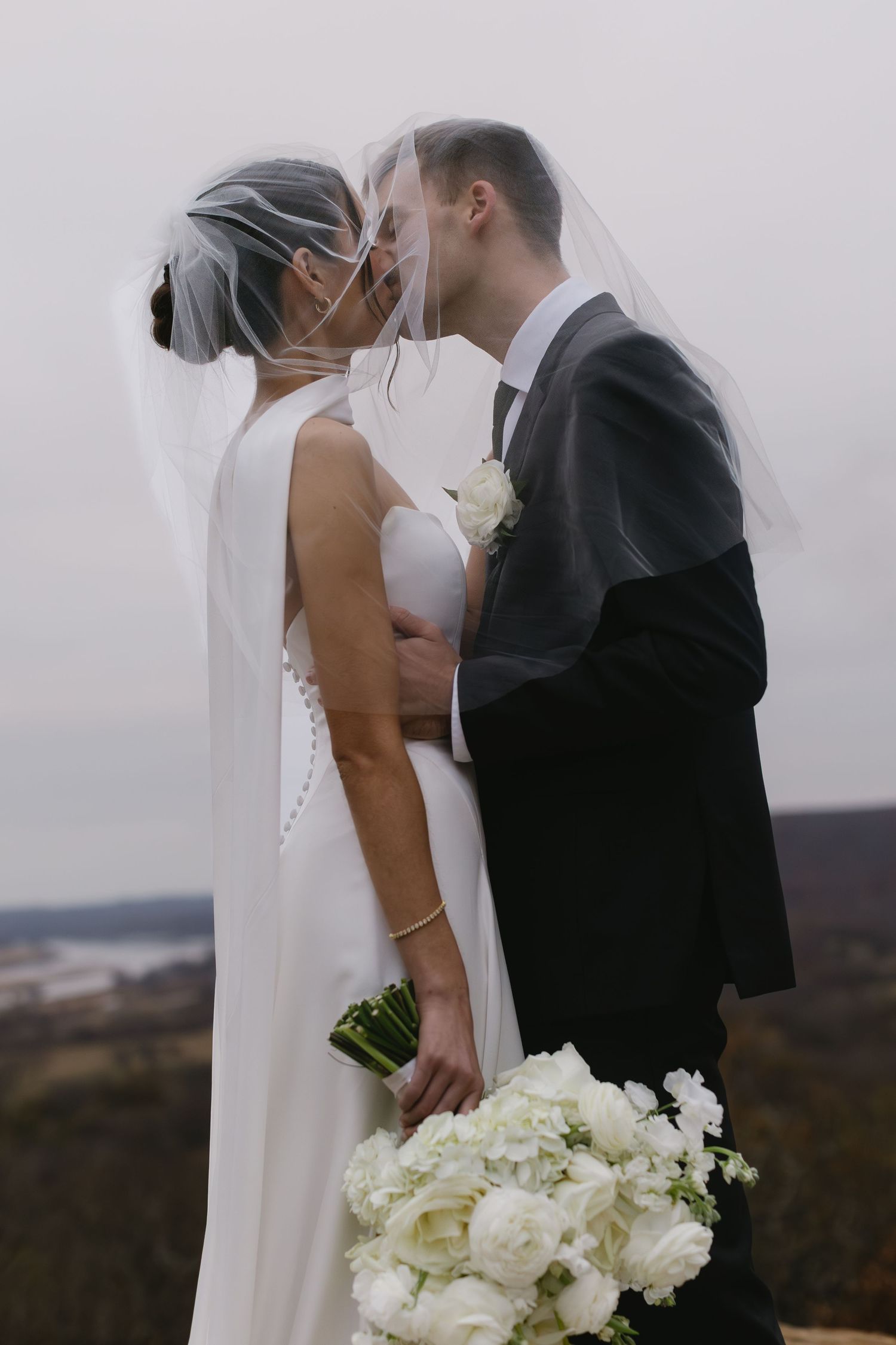 Couple shares intimate moment on mountaintop with flowing veil and white bouquet.