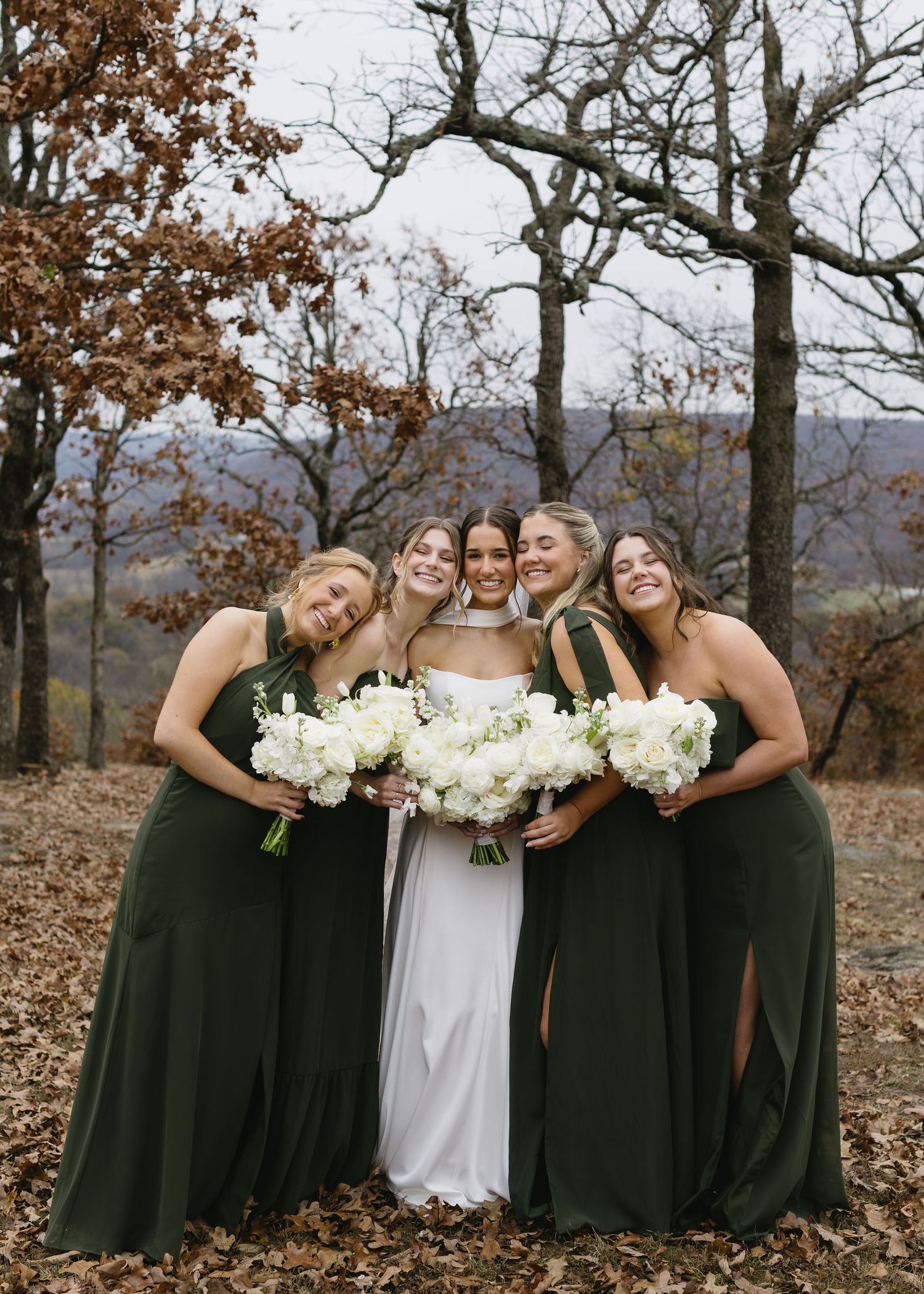A bride and bridesmaids in dark green dresses pose with white bouquets in a fall setting.