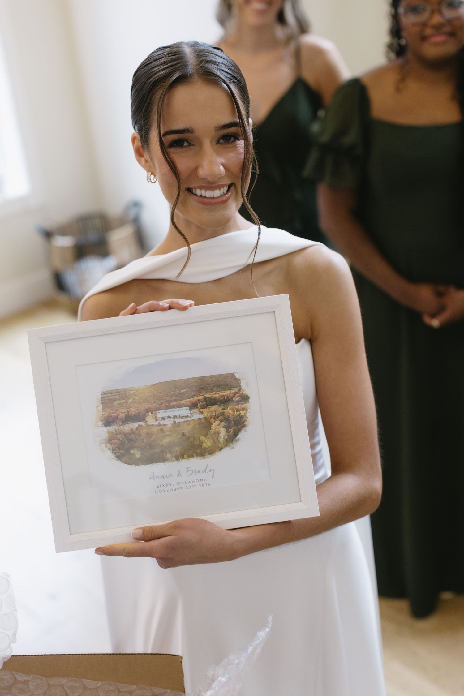 A bride in a white dress smiles while holding framed artwork during wedding preparations.