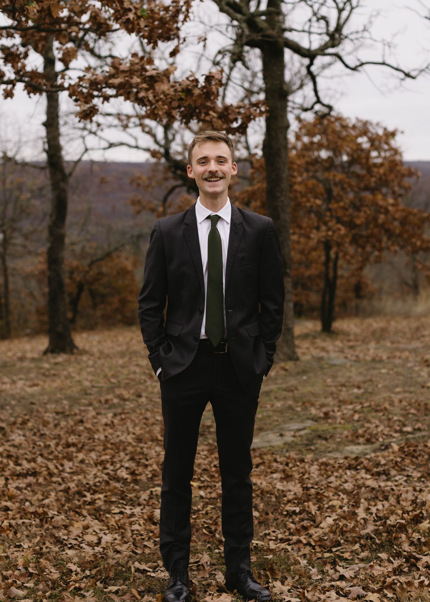 Groom in black suit and tie stands among fallen autumn leaves in a wooded outdoor setting.
