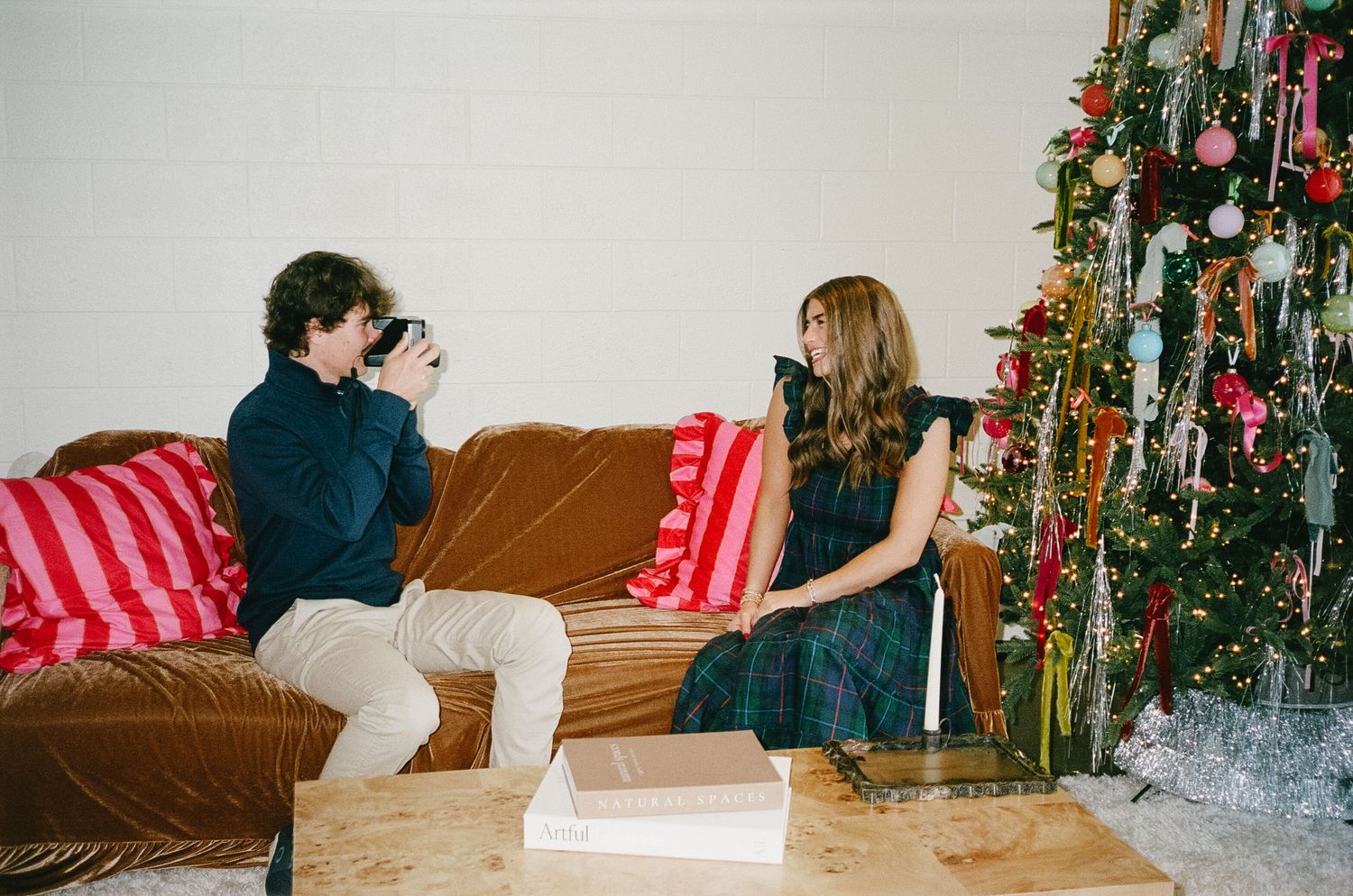 A couple sits on a couch near a decorated Christmas tree in a living room setting.