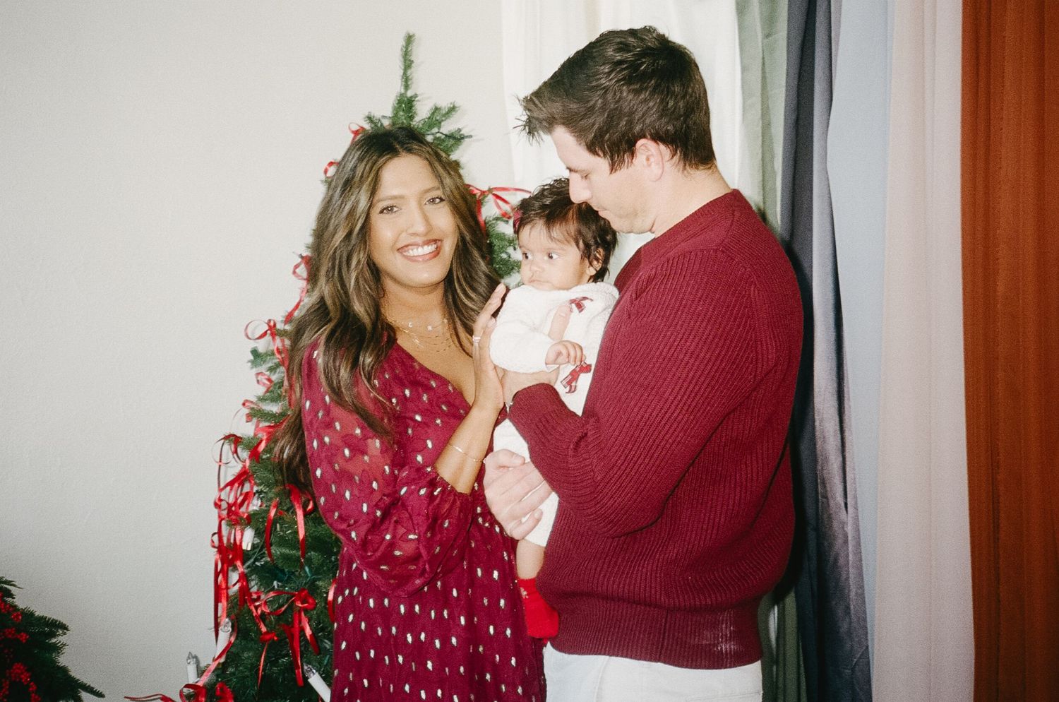 A couple in burgundy and red holiday attire share a romantic dance moment in a brightly lit room.