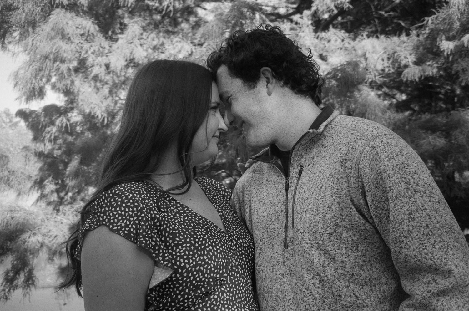 A romantic black and white portrait of a couple in casual clothing leaning close together outdoors.