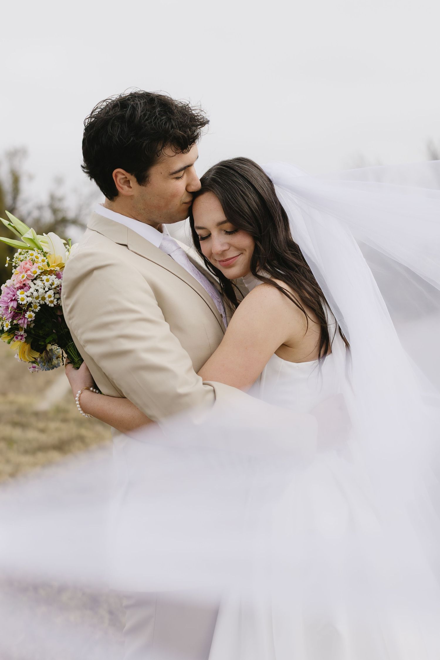 Romantic wedding couple embracing outdoors with flowing veil and light beige suit.