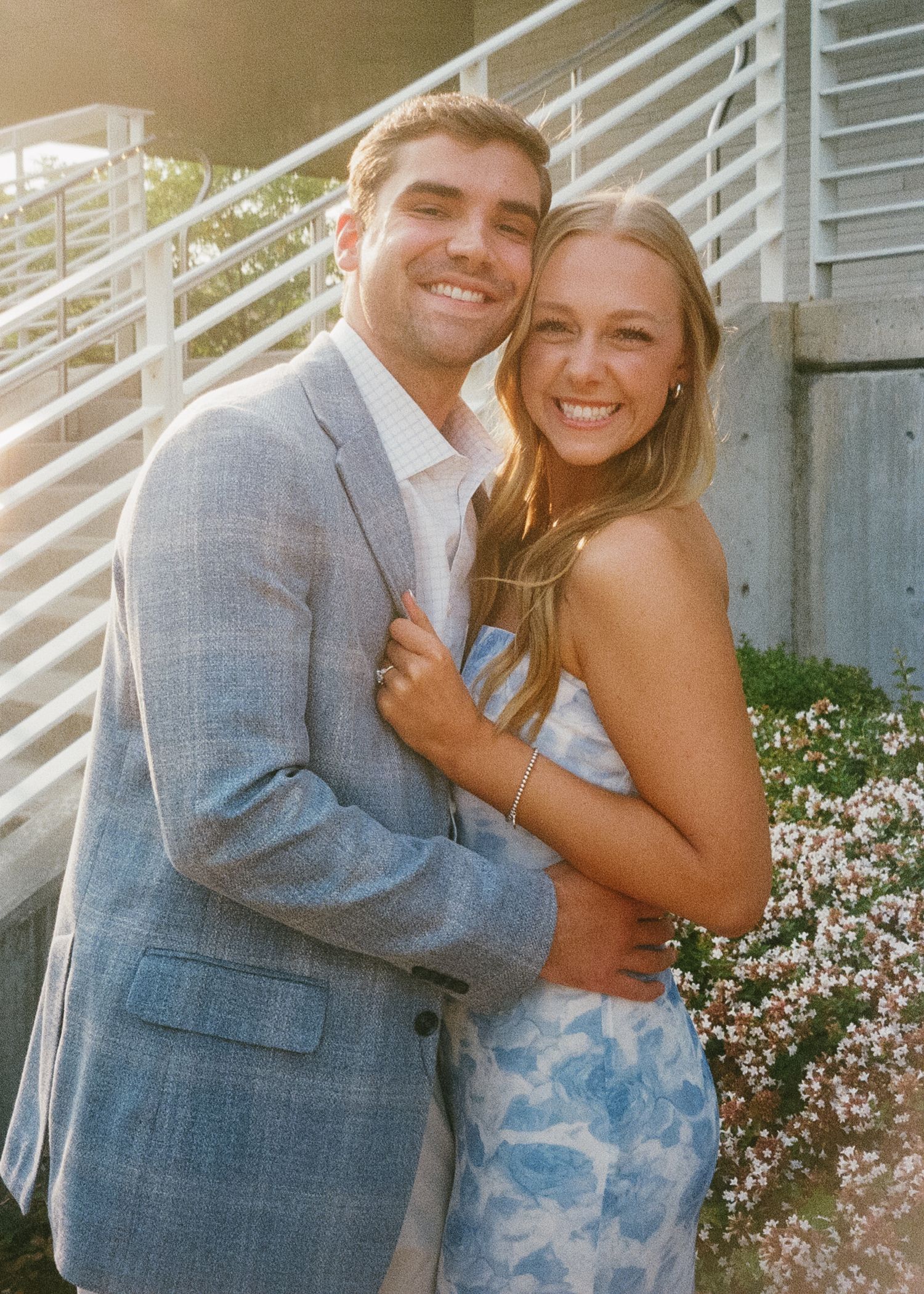A couple embraces and laughs together against a white railing with small pink flowers in the background.
