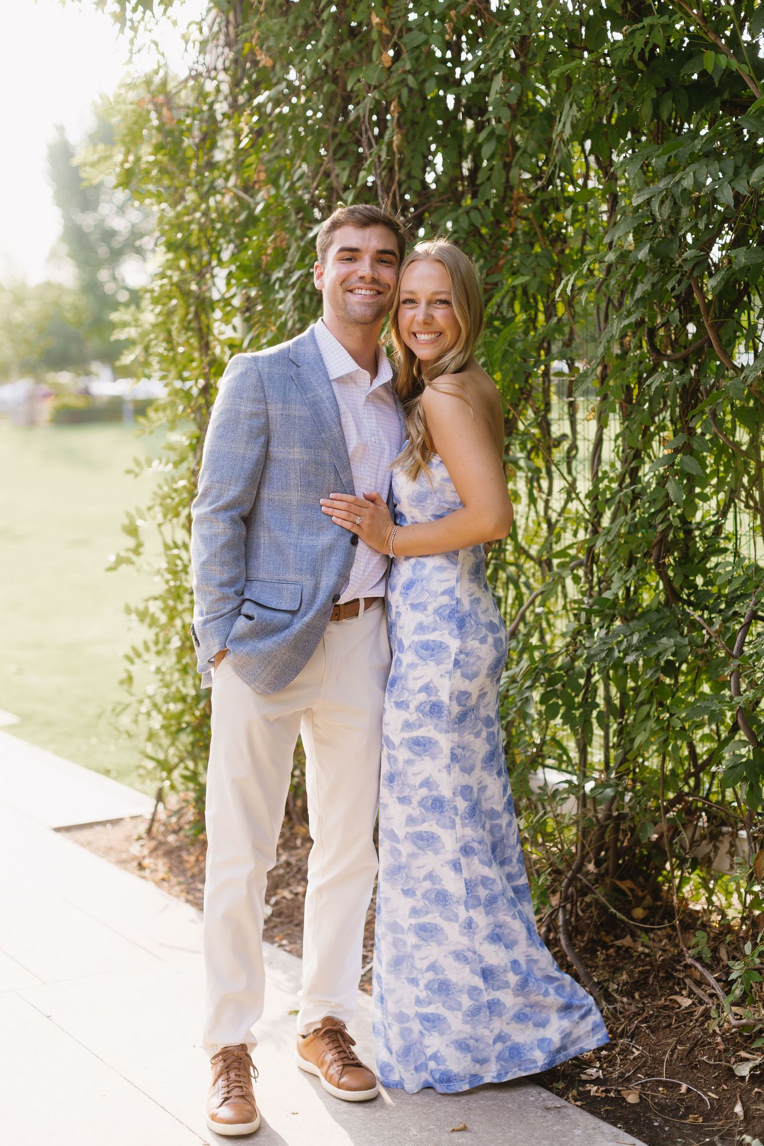A couple poses together on a garden path with willow trees and summer greenery in the background.