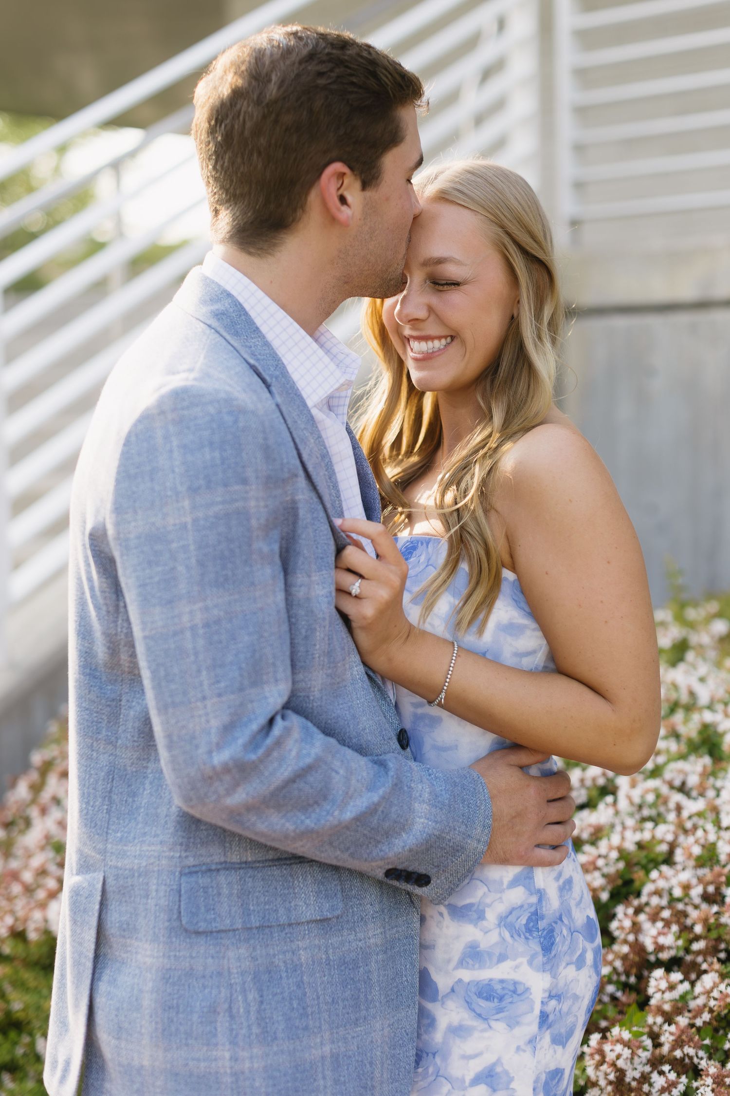 A couple in blue clothing share a romantic moment while laughing together against a white wall with flowering plants.