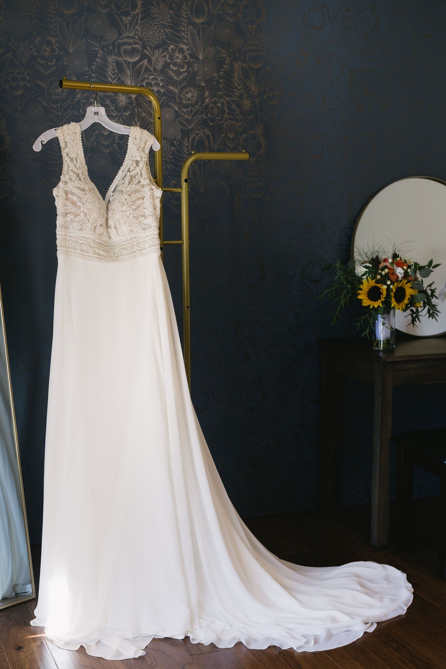 An elegant white wedding dress with lace bodice and flowing train displayed on a hanger against a dark patterned wall.