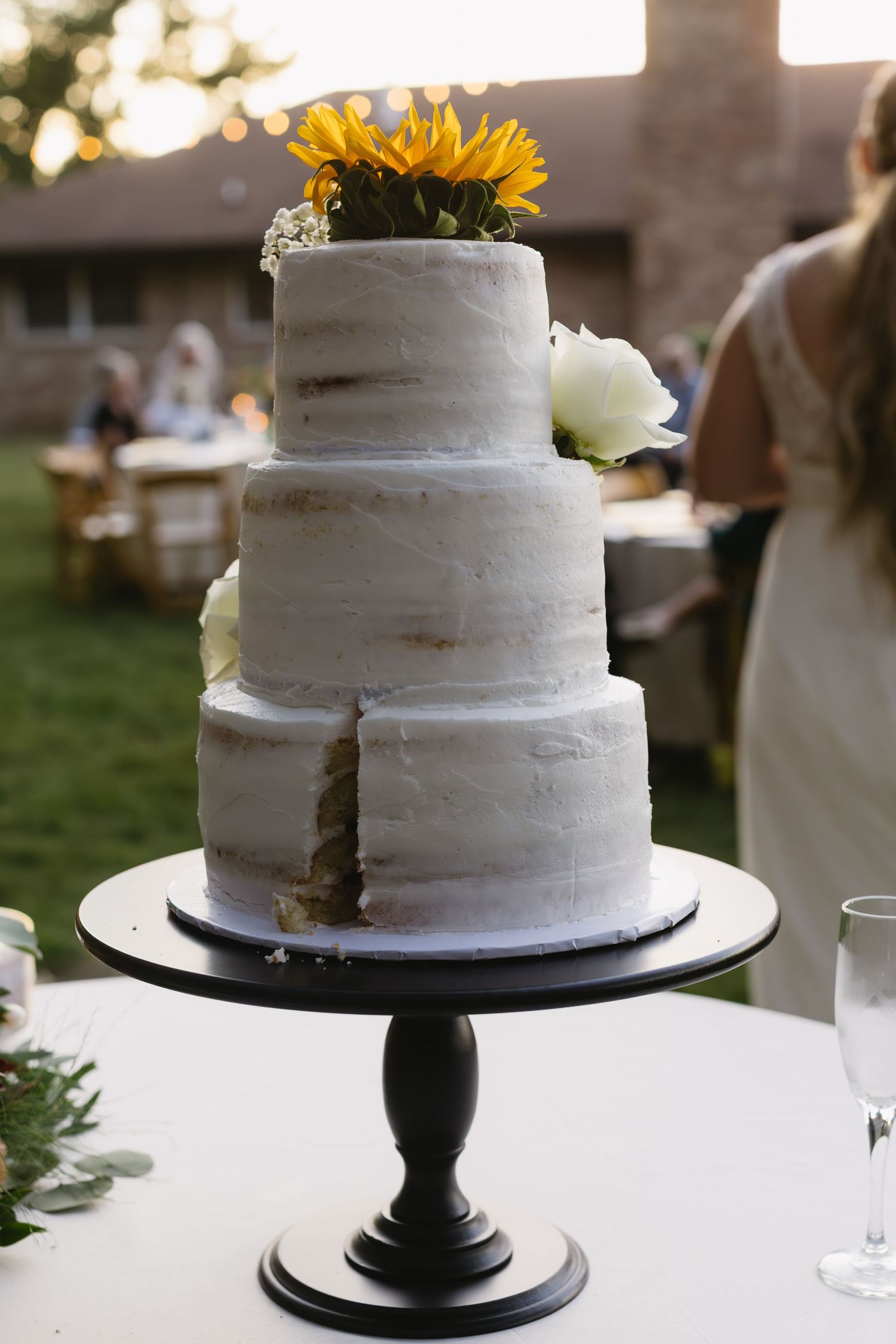 White tiered wedding cake decorated with sunflowers displayed on a black pedestal stand.