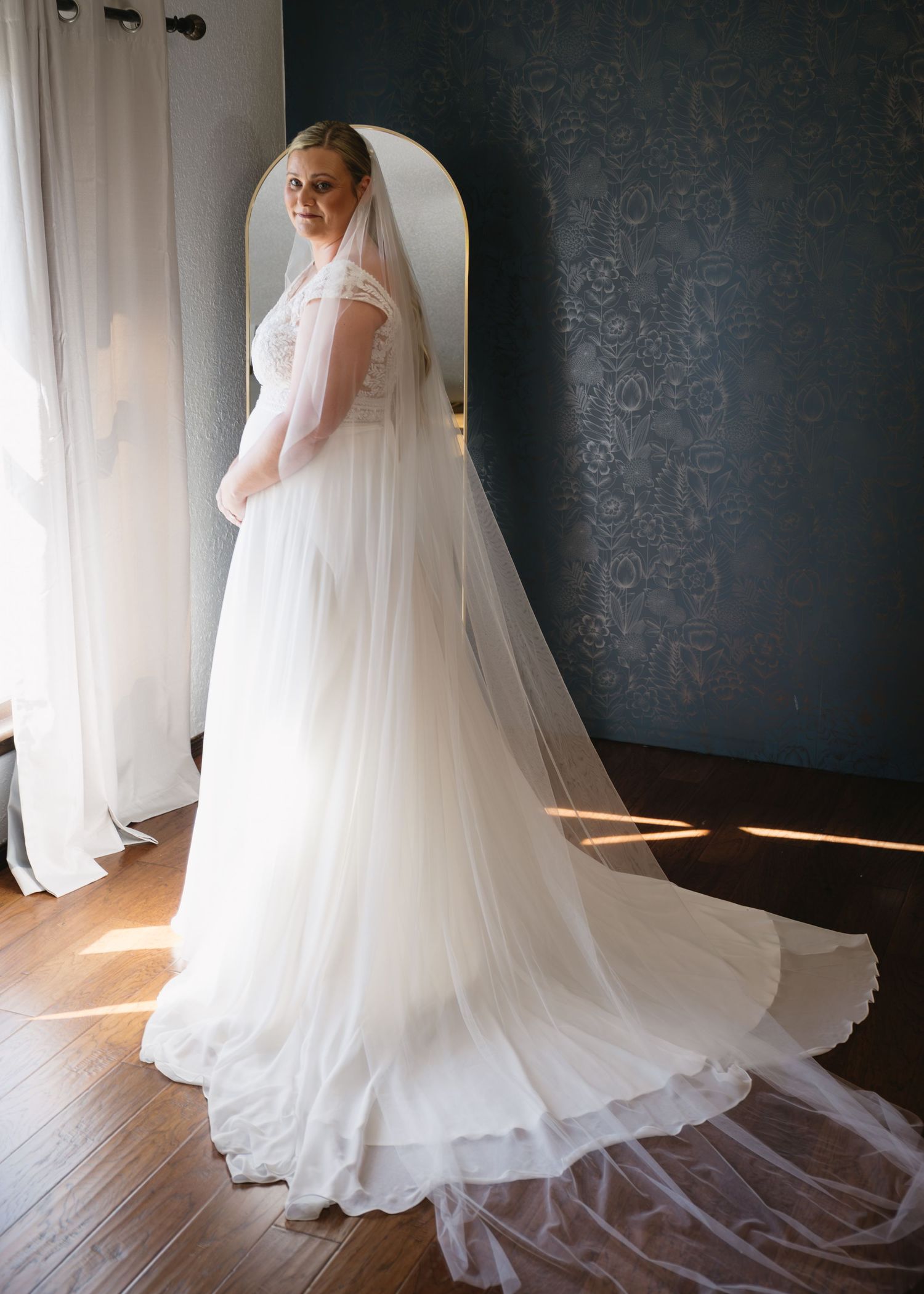 Ethereal wedding dress with long train and veil photographed against dark textured wall.