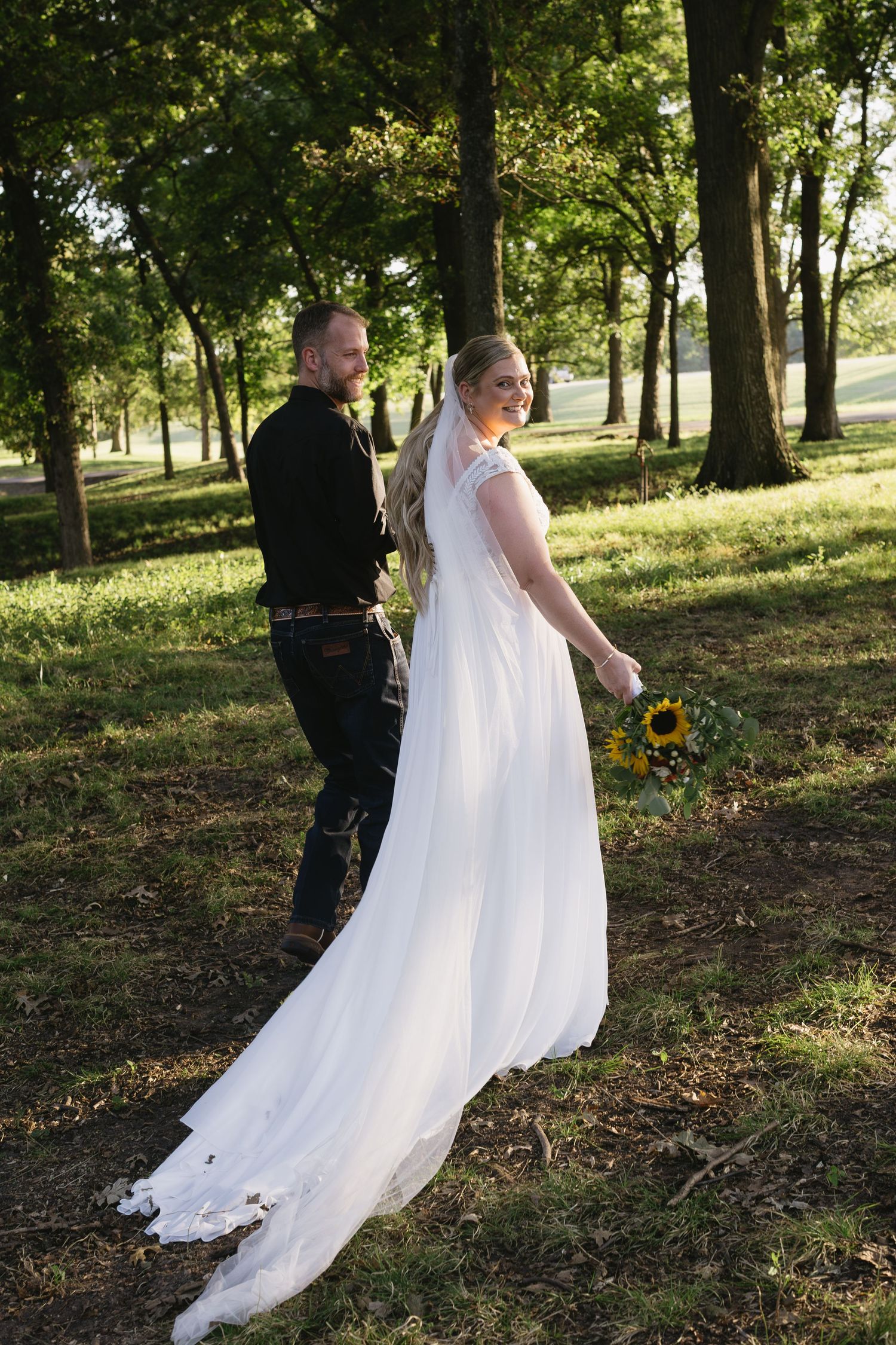 A couple walks through a wooded park setting with sunflowers and flowing white wedding dress.