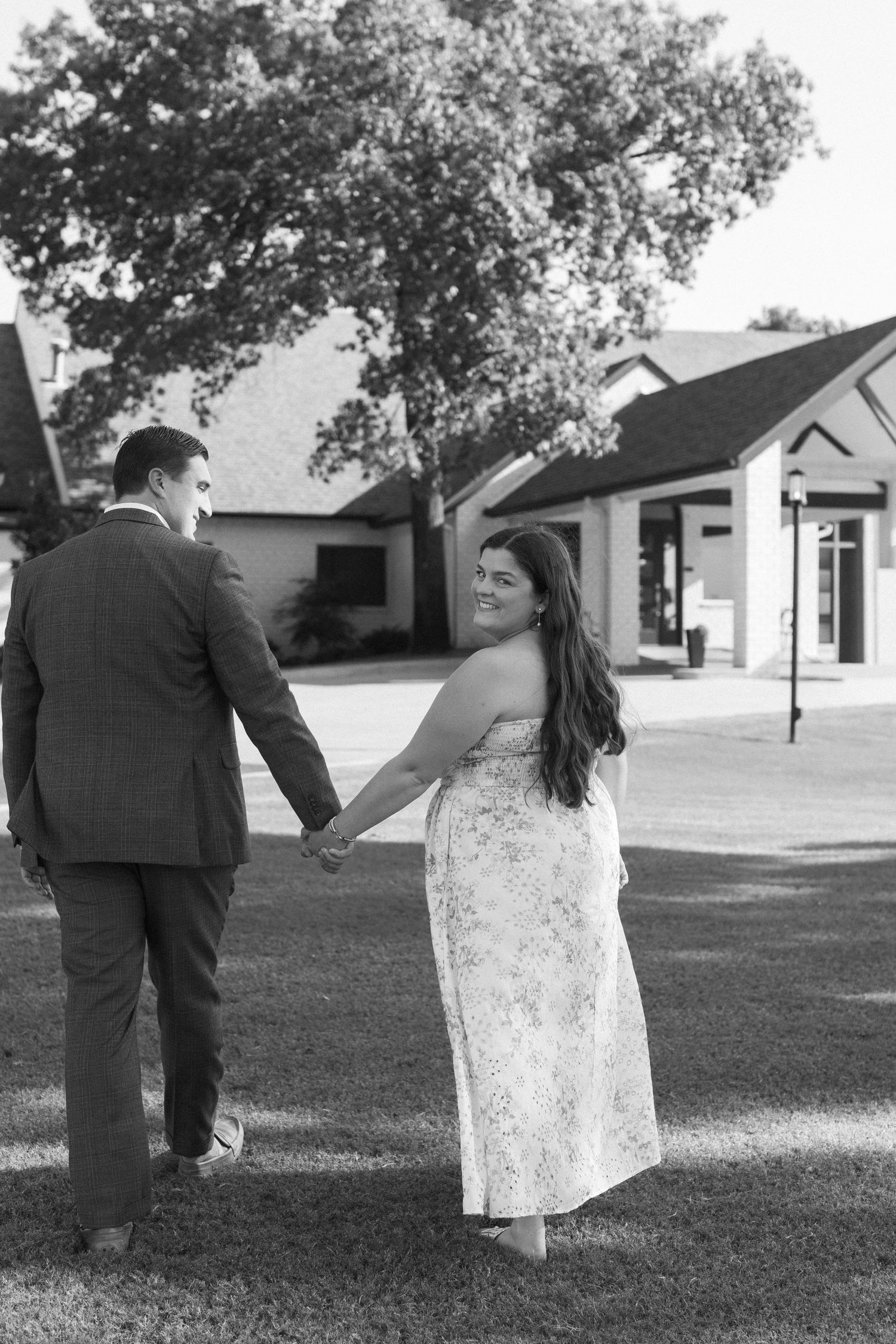 Black and white photo of a couple holding hands while walking on grass near buildings.