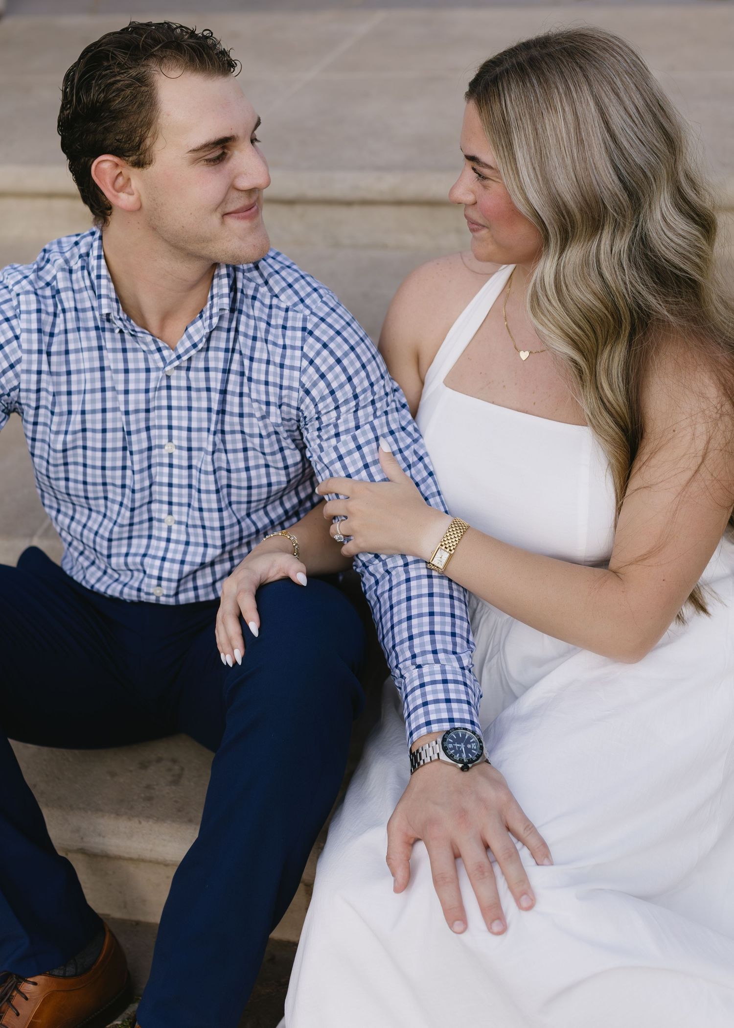 A couple poses together on steps wearing formal attire of white dress and checkered blue shirt.