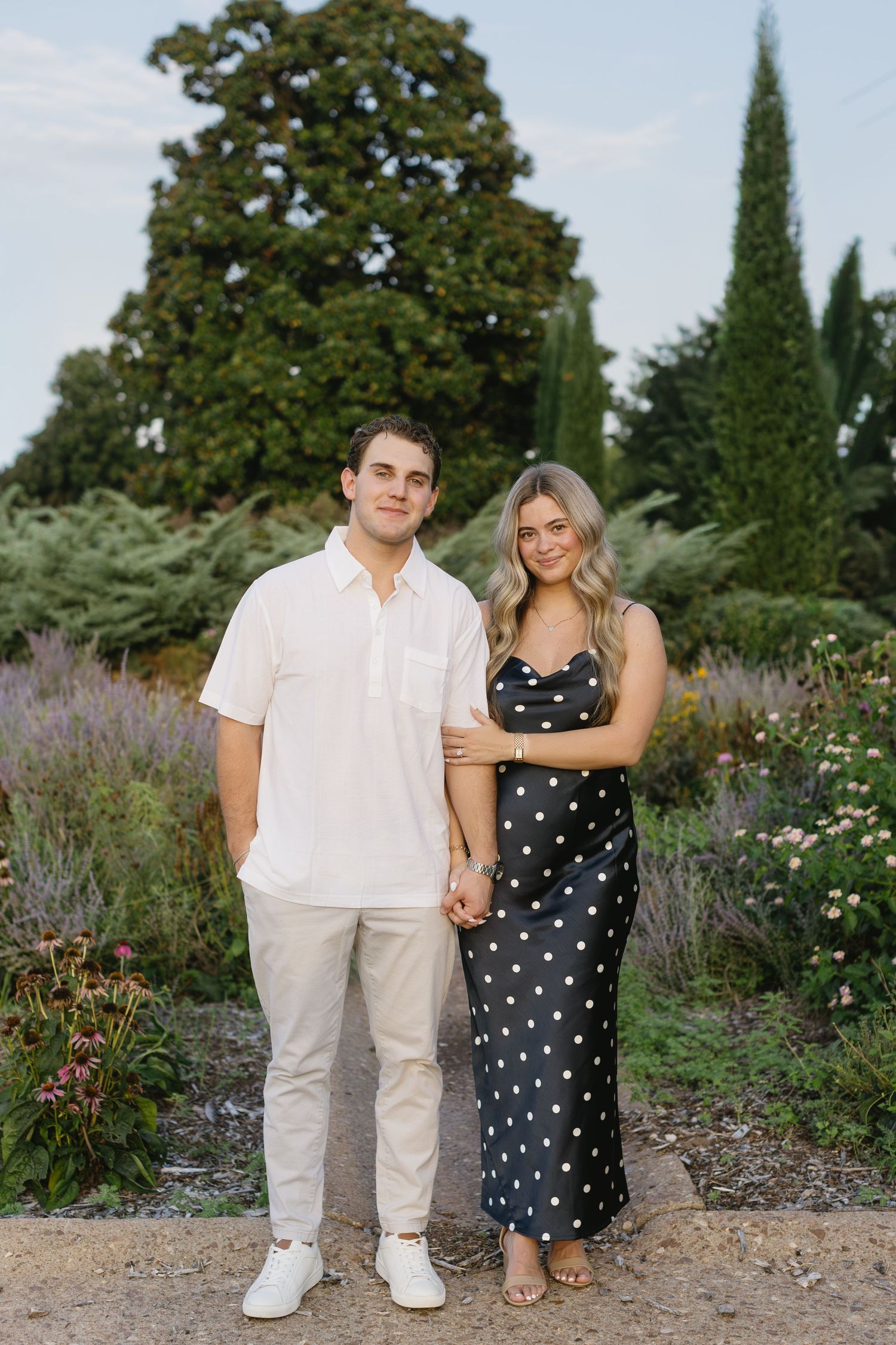 Romantic couple poses together in garden with white and polka dot outfits against flowering landscape.