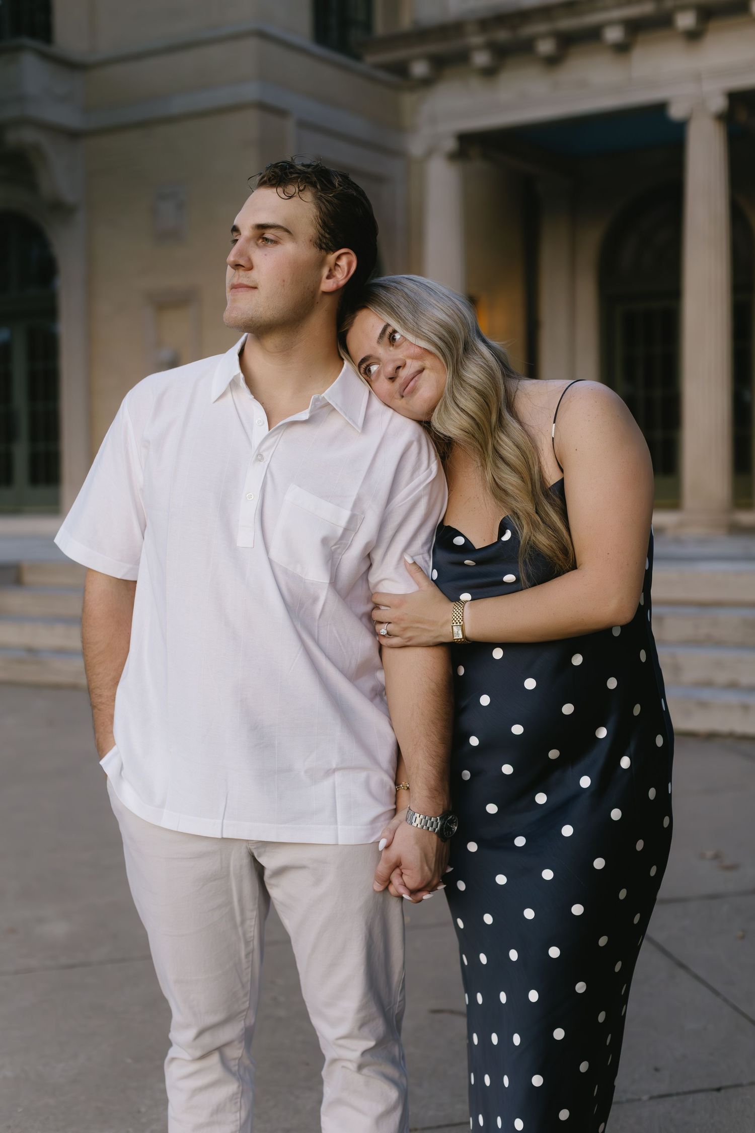 Couple poses against architectural backdrop, woman in polka dot dress leans affectionately on man in white shirt.