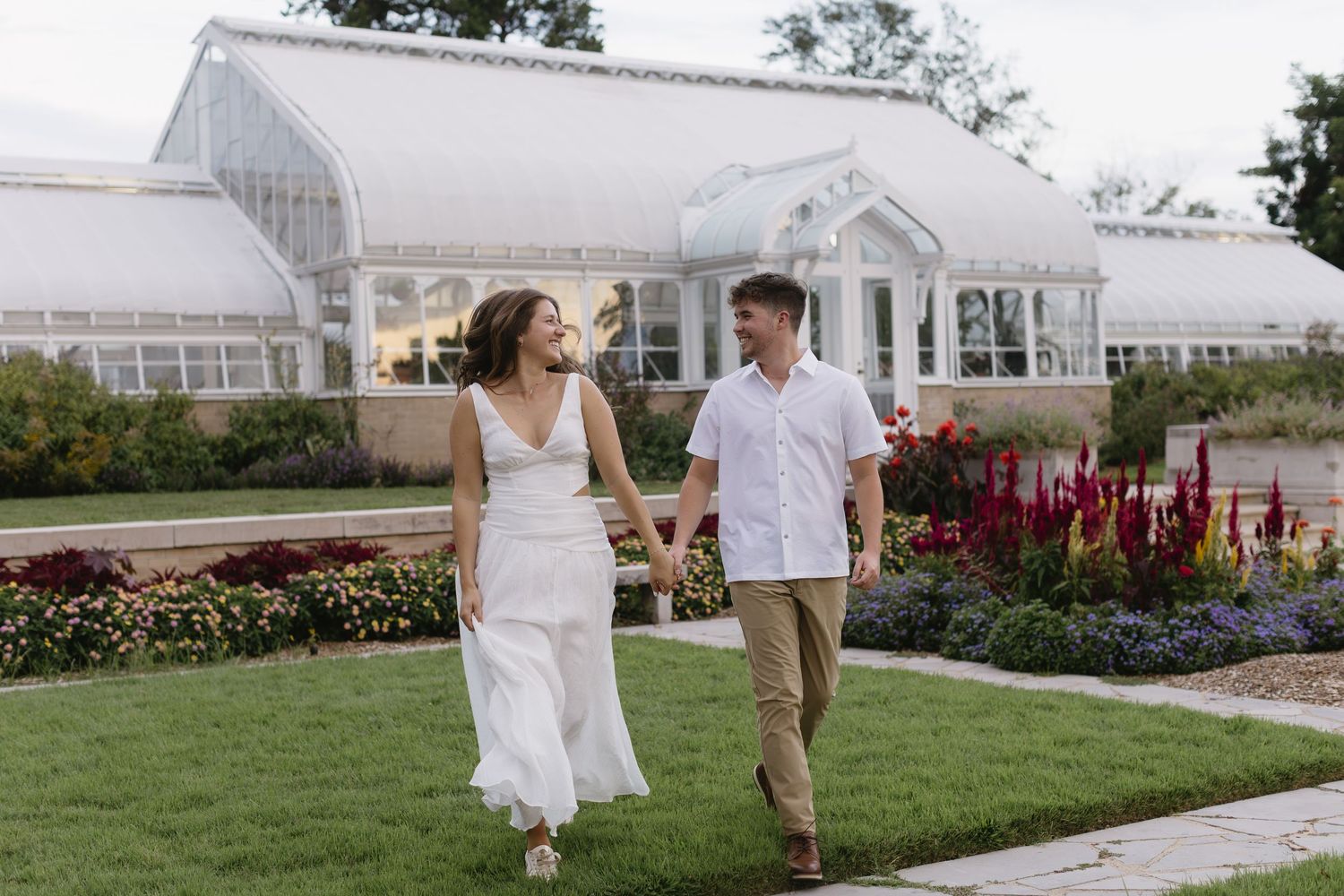 A couple in white attire poses playfully in front of a grand white building with gardens.
